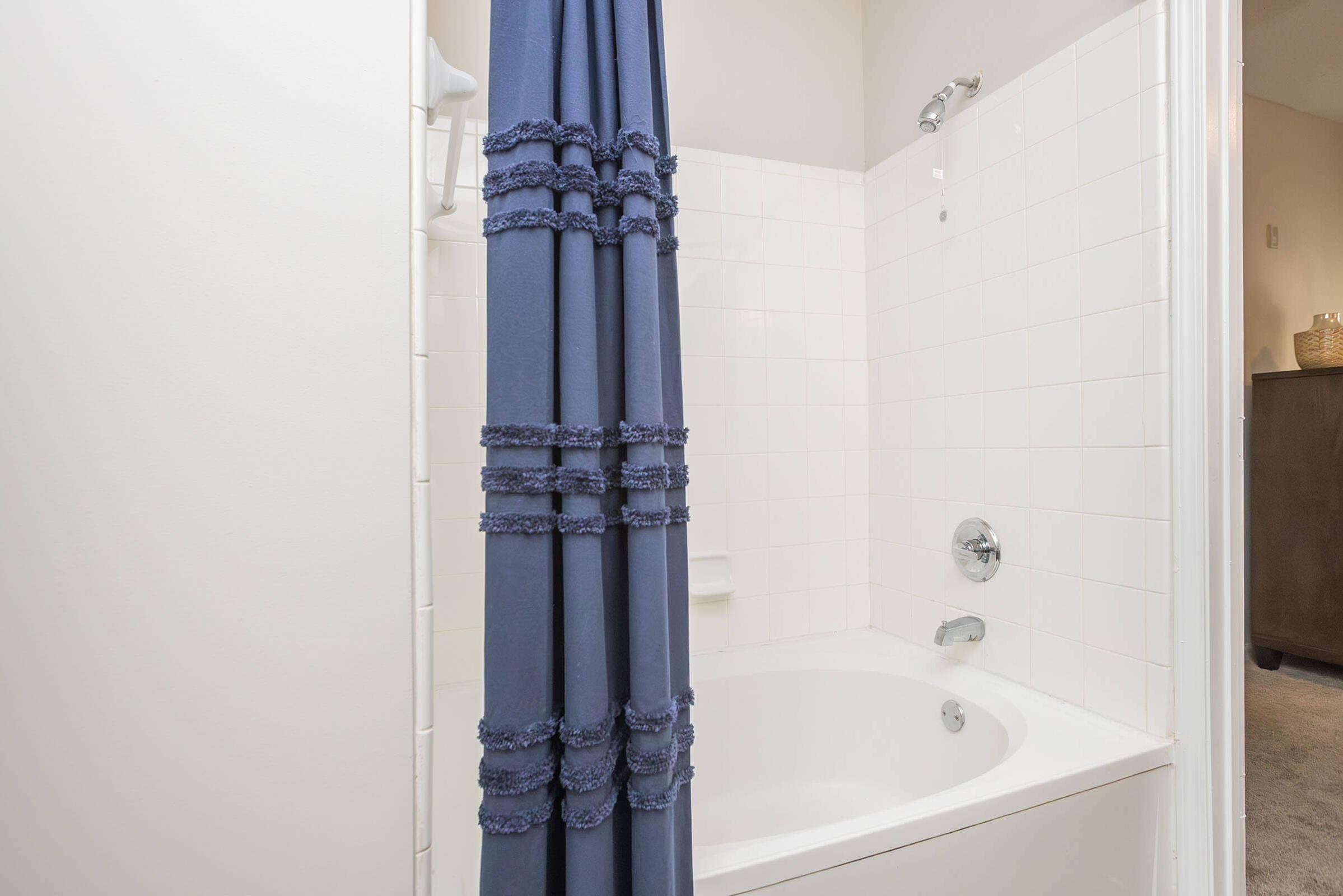 A modern bathroom featuring a bathtub with a shower. The bathtub is partially covered by a dark blue shower curtain adorned with ruffles. The tiled walls are white, and fixtures like a showerhead and faucet are visible. A framed mirror is faintly seen in the background.