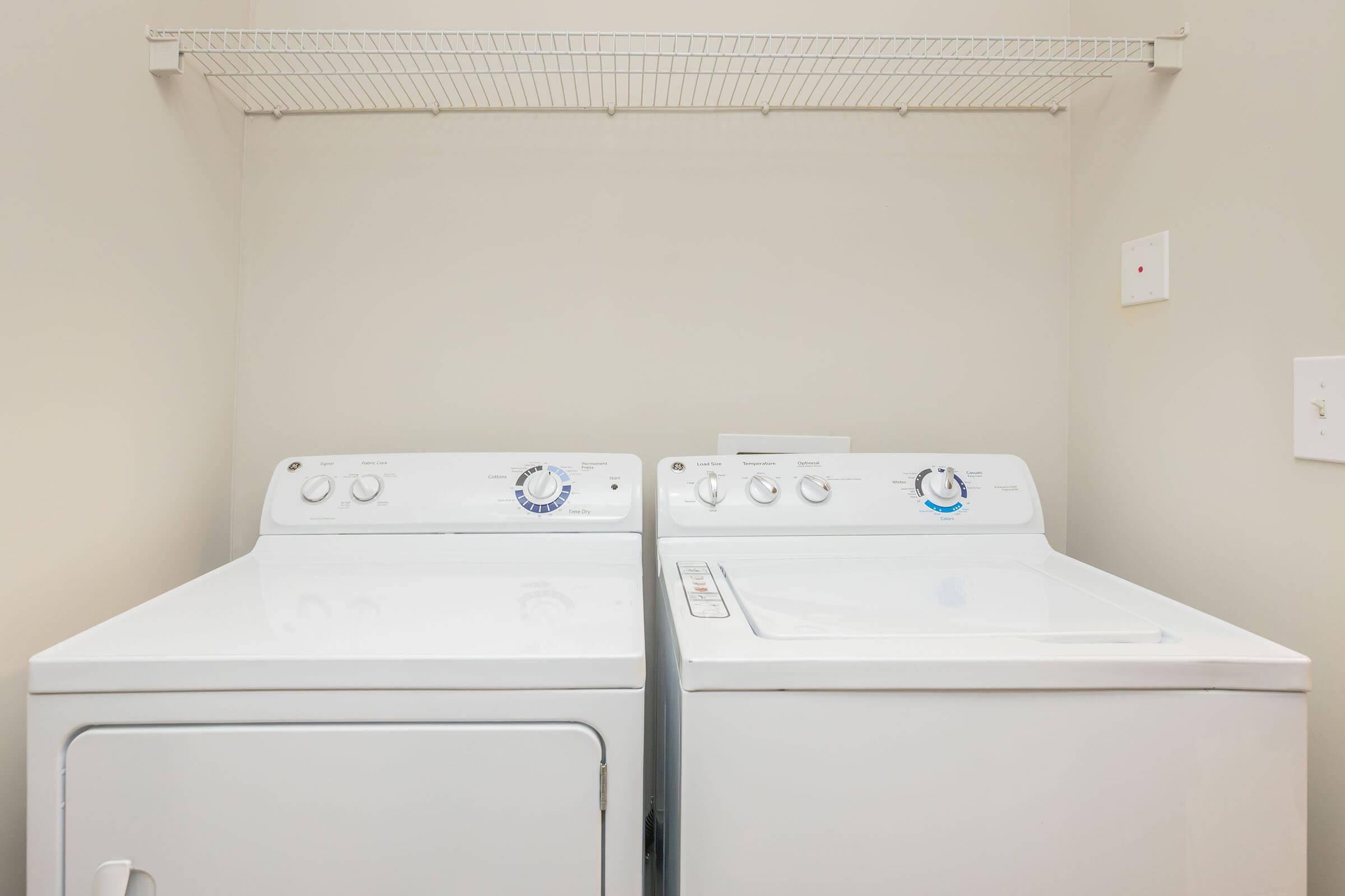 A laundry room featuring a stacked white washing machine and dryer, placed side by side against a light-colored wall. Above them, there is a wire shelf for storage. The appliances have control dials and buttons visible on their front panels.