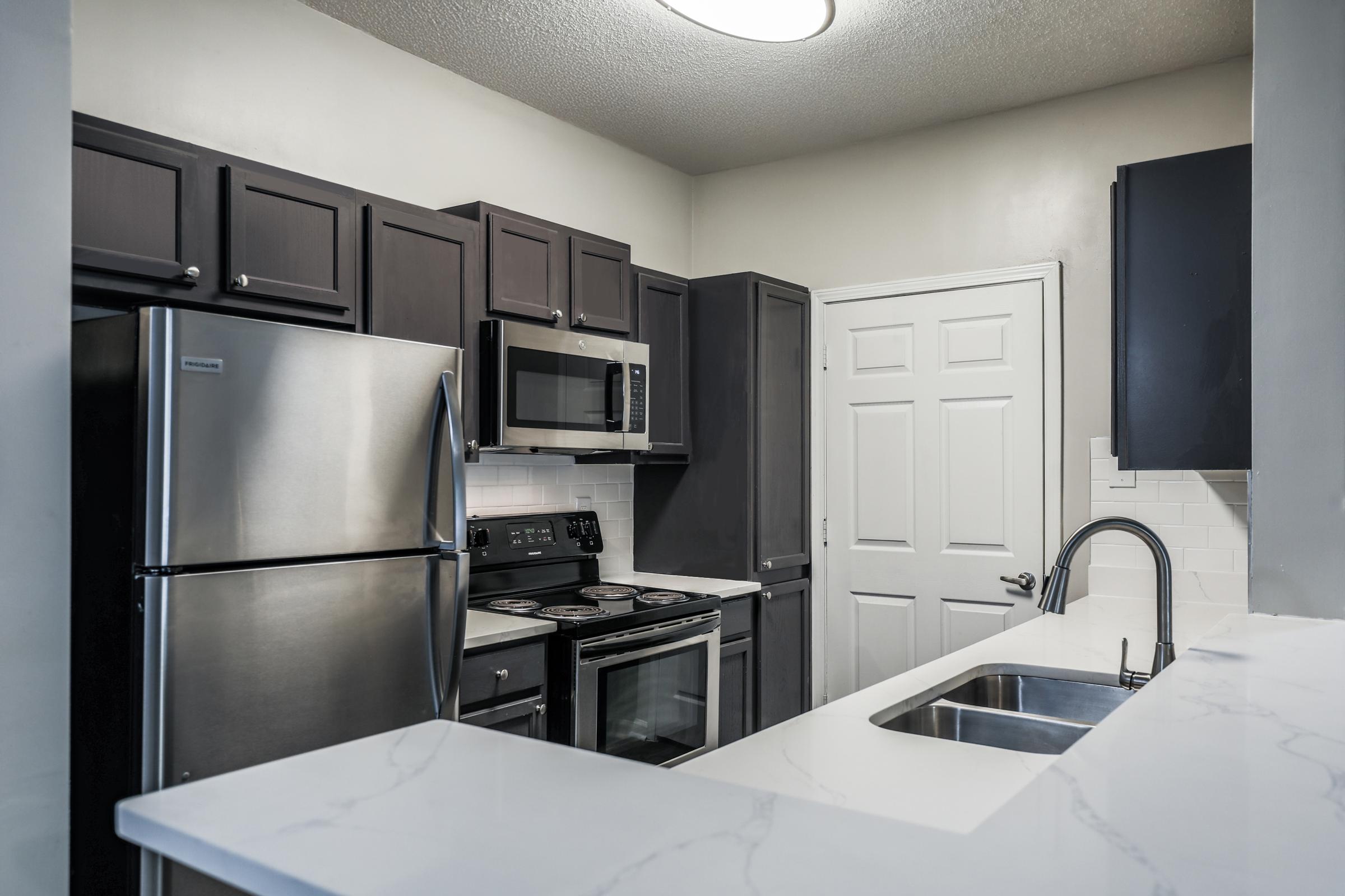 Modern kitchen featuring dark cabinetry, stainless steel appliances, and a white countertop. The kitchen includes a refrigerator, microwave, oven, and a sink area with a faucet. A white door leads to another room, and the space is illuminated by overhead lighting.