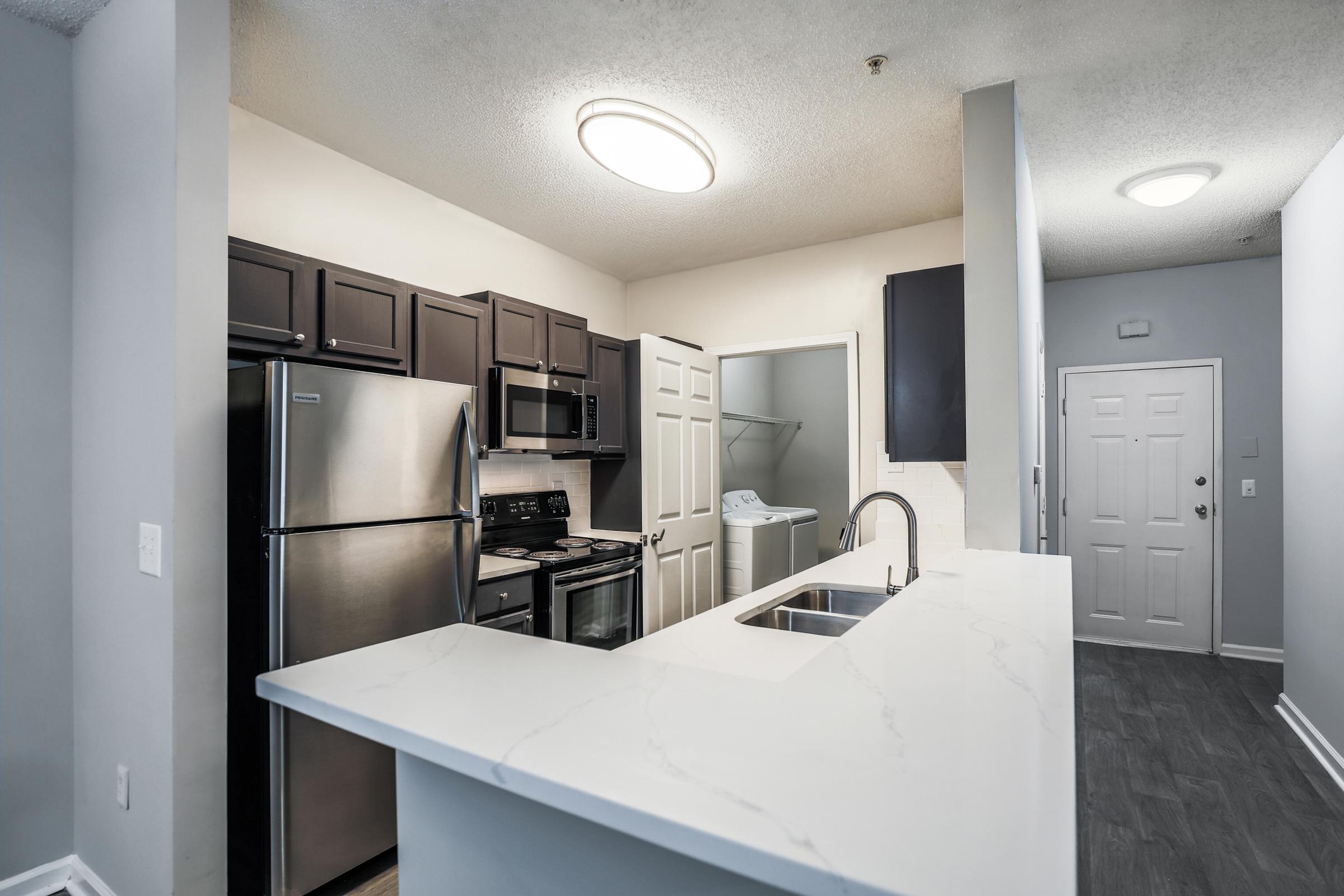 Modern kitchen featuring dark wood cabinets, stainless steel appliances including a refrigerator, stove, and dishwasher. A white countertop with a sink is visible in the foreground. The layout includes a doorway leading to a laundry area and a front door in the background. Soft lighting fills the space.