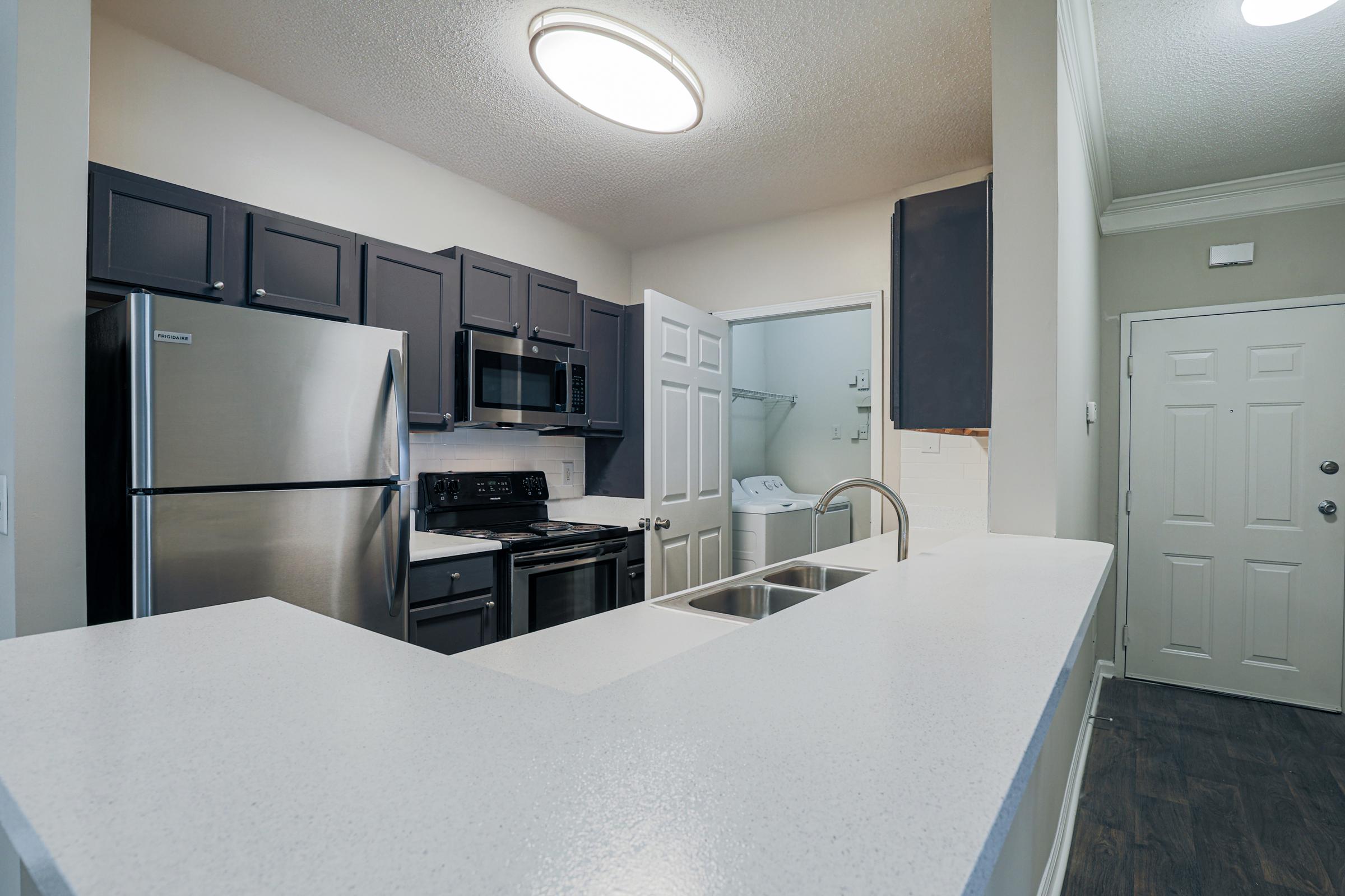 Modern kitchen featuring stainless steel appliances, including a refrigerator and microwave, dark cabinetry, and a breakfast bar. A door leads into a laundry area, while another door provides access to the entrance of the home. The space is well-lit with a ceiling light fixture.
