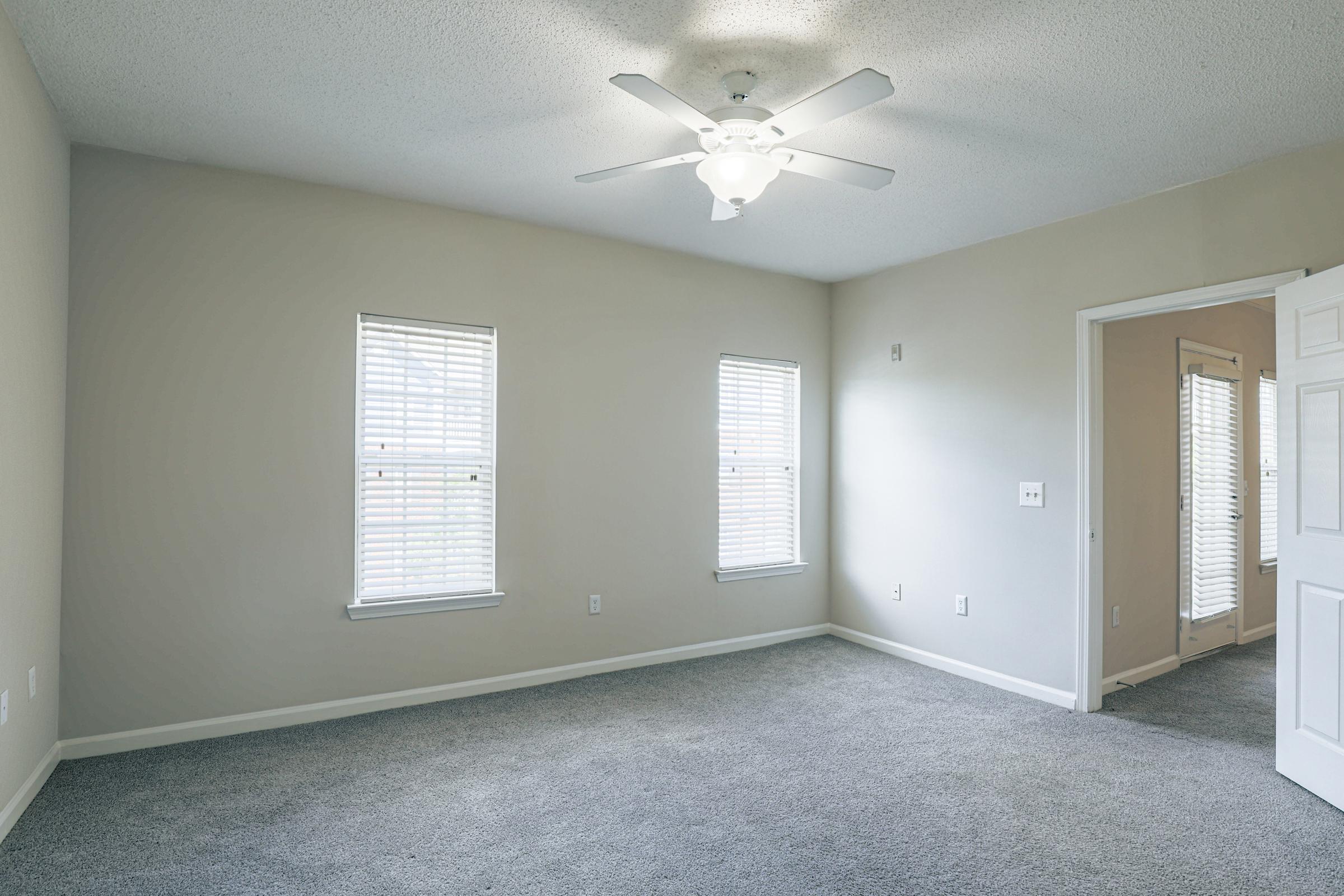 A bright, empty room with light gray carpet, white walls, and a ceiling fan. There are three windows with white blinds allowing natural light to enter. One wall has a door leading to another space, and the room appears spacious and ready for furnishing.