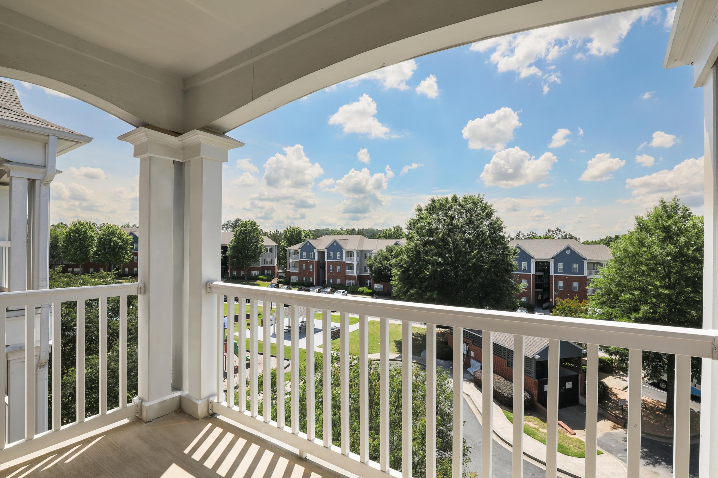 A spacious balcony view overlooking a vibrant neighborhood. The scene features multiple buildings with green lawns, trees, and a clear blue sky adorned with fluffy white clouds, creating a serene outdoor atmosphere.