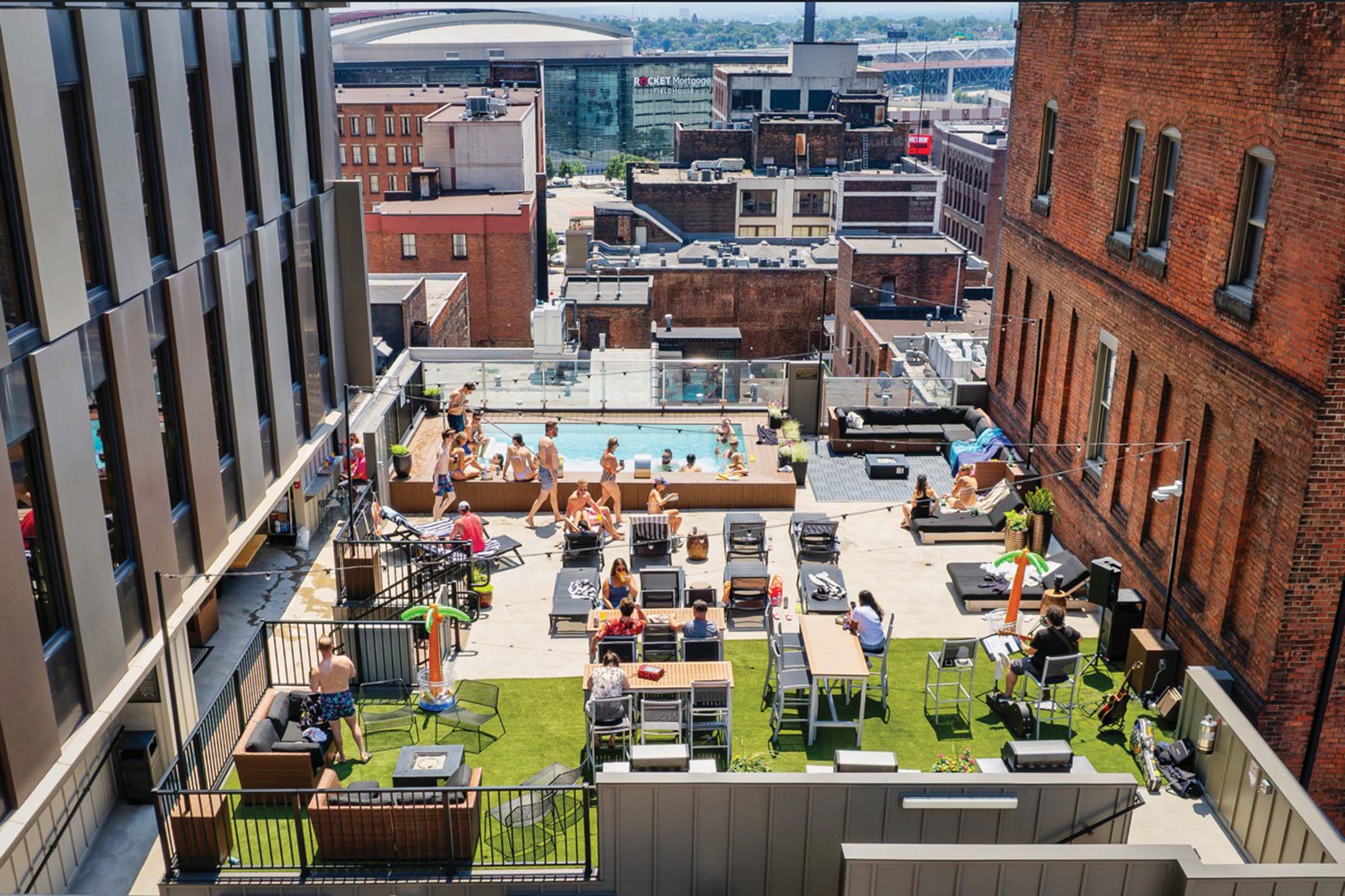 A rooftop pool area in an urban setting, featuring people lounging on sunbeds, relaxing on sofas, and enjoying the sun on a green lawn. Surrounding buildings create a cityscape backdrop, with some people swimming in the pool. Bright, sunny ambiance with a lively atmosphere.