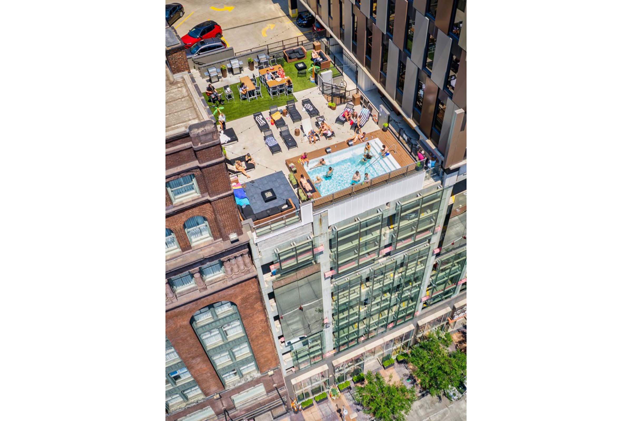 Aerial view of a rooftop pool area featuring sun loungers and a small pool, surrounded by greenery and seated spaces. Below, a busy city street is visible with parked cars and people walking by. The building combines modern glass architecture with a historic brick structure.
