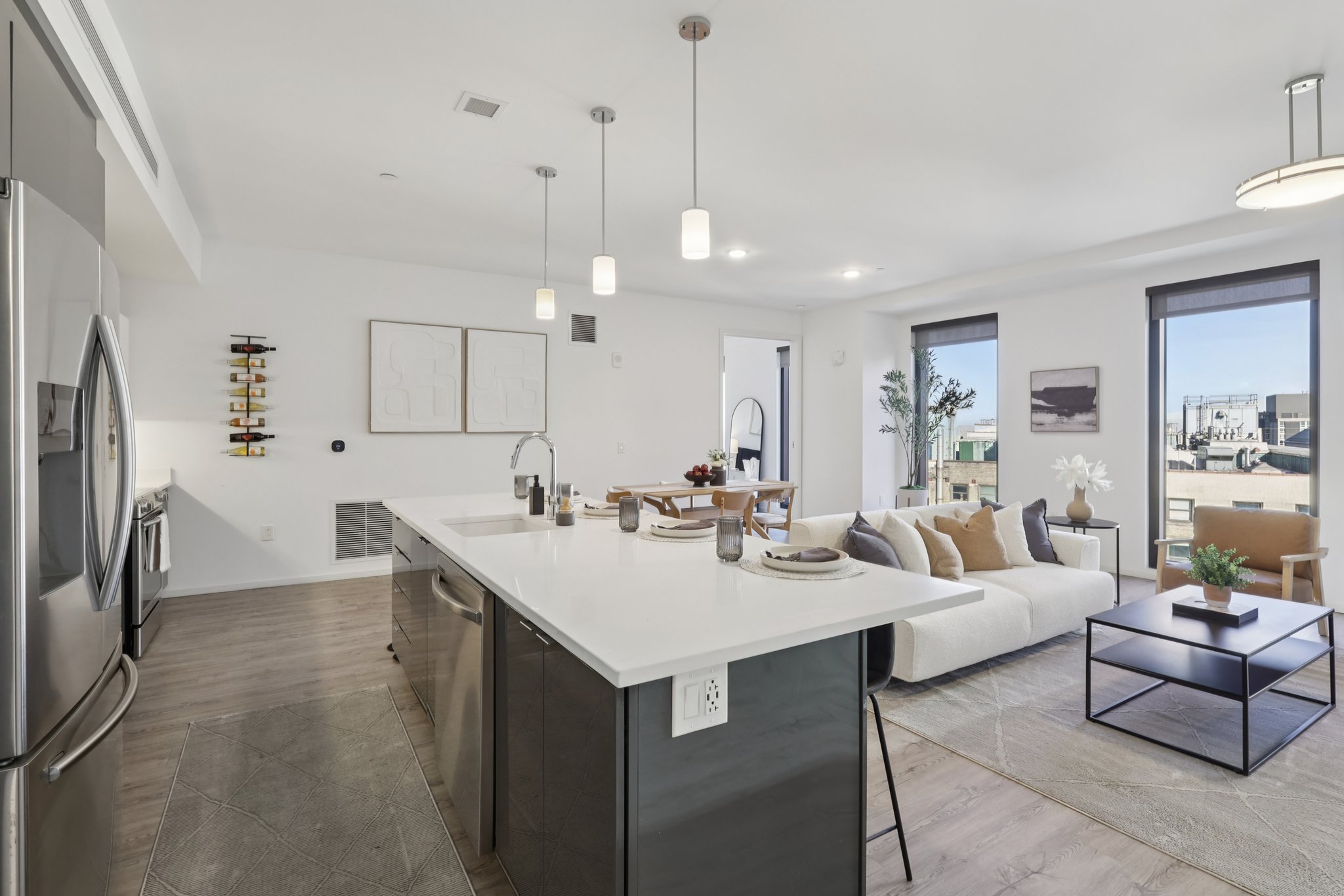A modern open-concept living space featuring a sleek kitchen with an island, stainless steel appliances, and a dining area. The living room has a cozy sofa and coffee table, with large windows providing natural light and a view of the outdoors. Neutral color palette with stylish decor.
