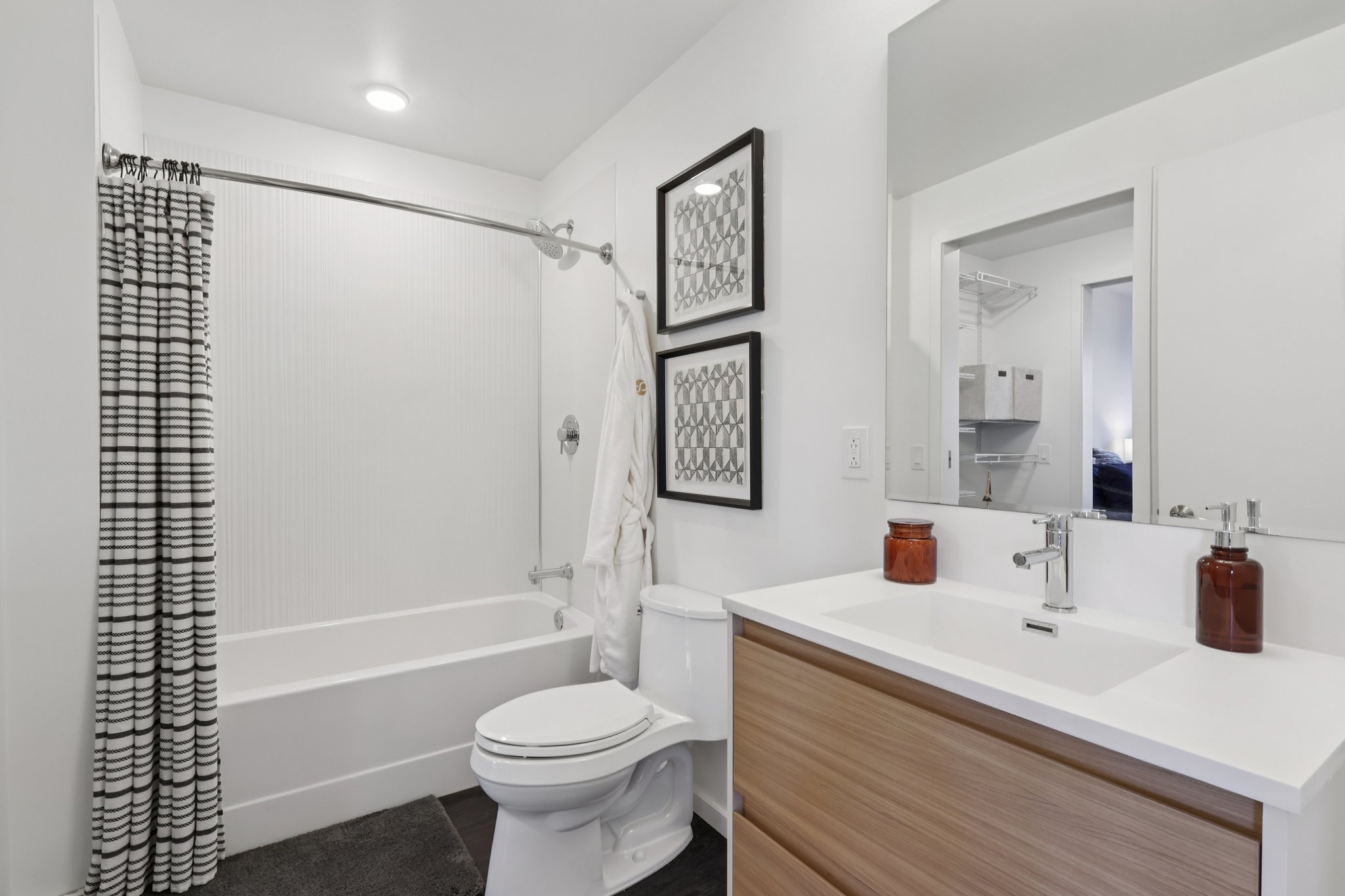 A modern bathroom featuring a shower-tub combo with a striped shower curtain, a clean white wall, a sleek wooden vanity with a sink, and a large mirror. Decorative black and white art frames hang on the wall, and a plush white robe hangs on a hook. The space is bright and well-lit.