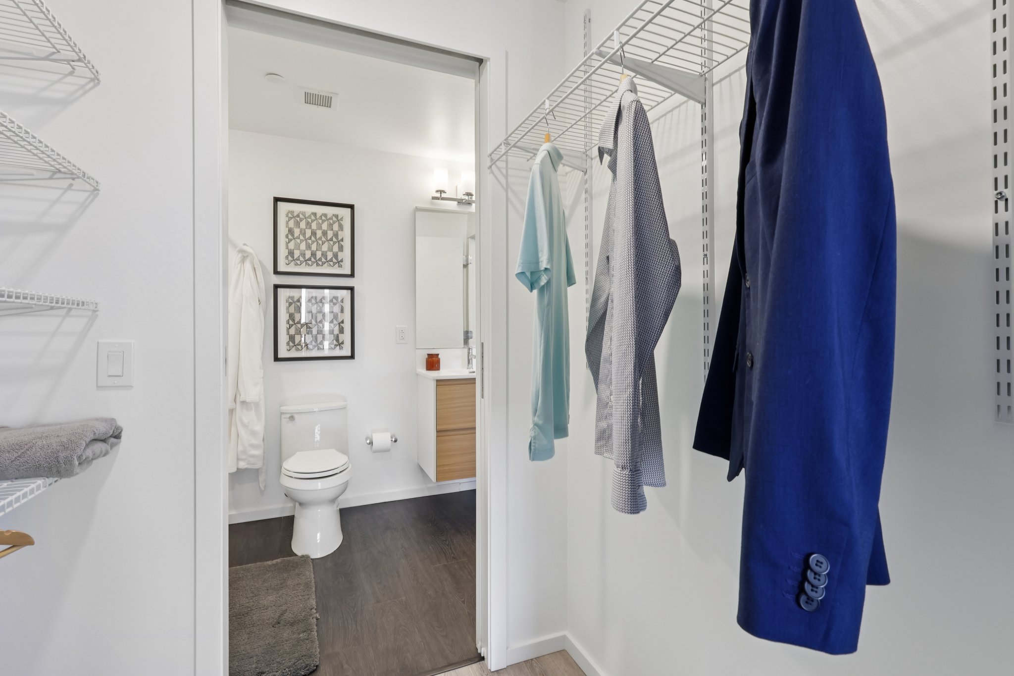 A modern bathroom view featuring a toilet, a vanity with a mirror, and light-colored walls. There are metal wire shelves with clothes and towels hanging nearby, creating a clean and organized look. Natural light illuminates the space, giving it a bright and airy feel.