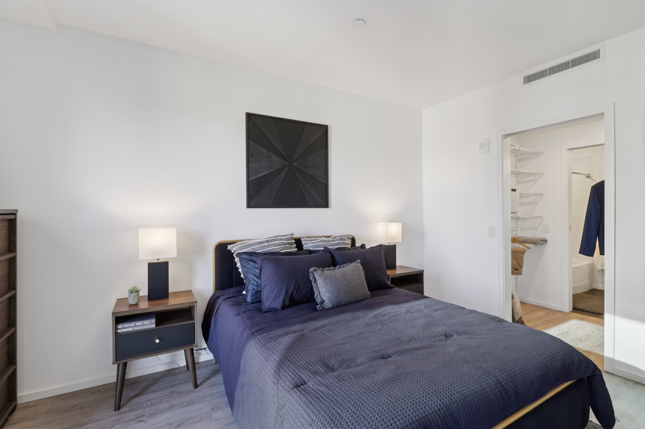 A modern bedroom featuring a bed with dark blue bedding, two bedside lamps, and a geometric black wall art piece. The room has light-colored walls, a dresser, and a closet with open doors showing shelves. Natural light is present, creating a bright and inviting atmosphere.