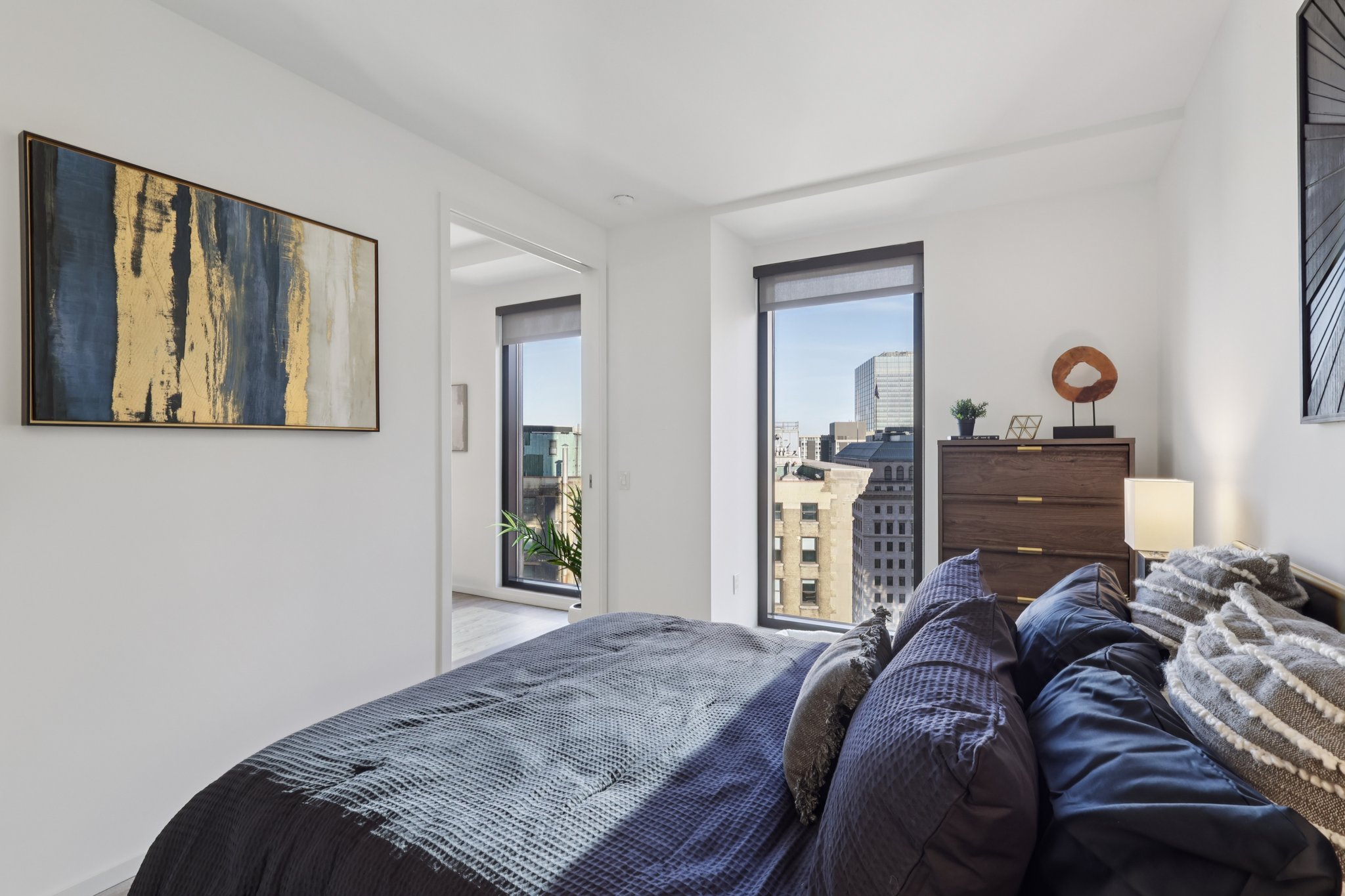 A modern bedroom featuring a dark blue bedspread with decorative pillows. The walls are white, and a large piece of abstract art in gold and blue hangs above a wooden dresser. Natural light streams in through large windows, revealing a city skyline view. A small plant is visible on the balcony outside.