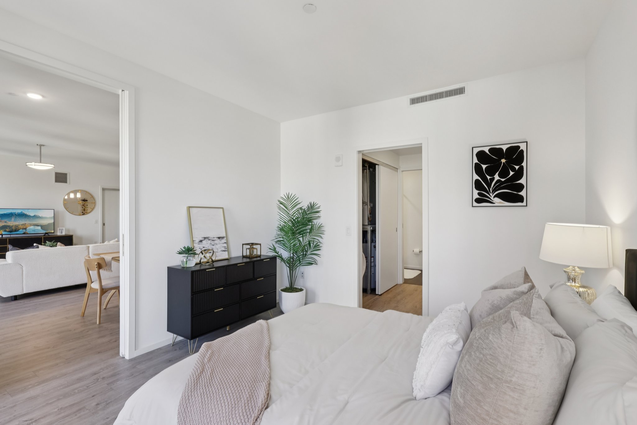 Cozy bedroom featuring a modern design with a bed adorned in soft pillows and bedding. A sleek black dresser with a decorative plant is positioned beside the bed. The room is bright, with a doorway leading to another area of the home, showcasing a minimalistic aesthetic and natural light filtering through.