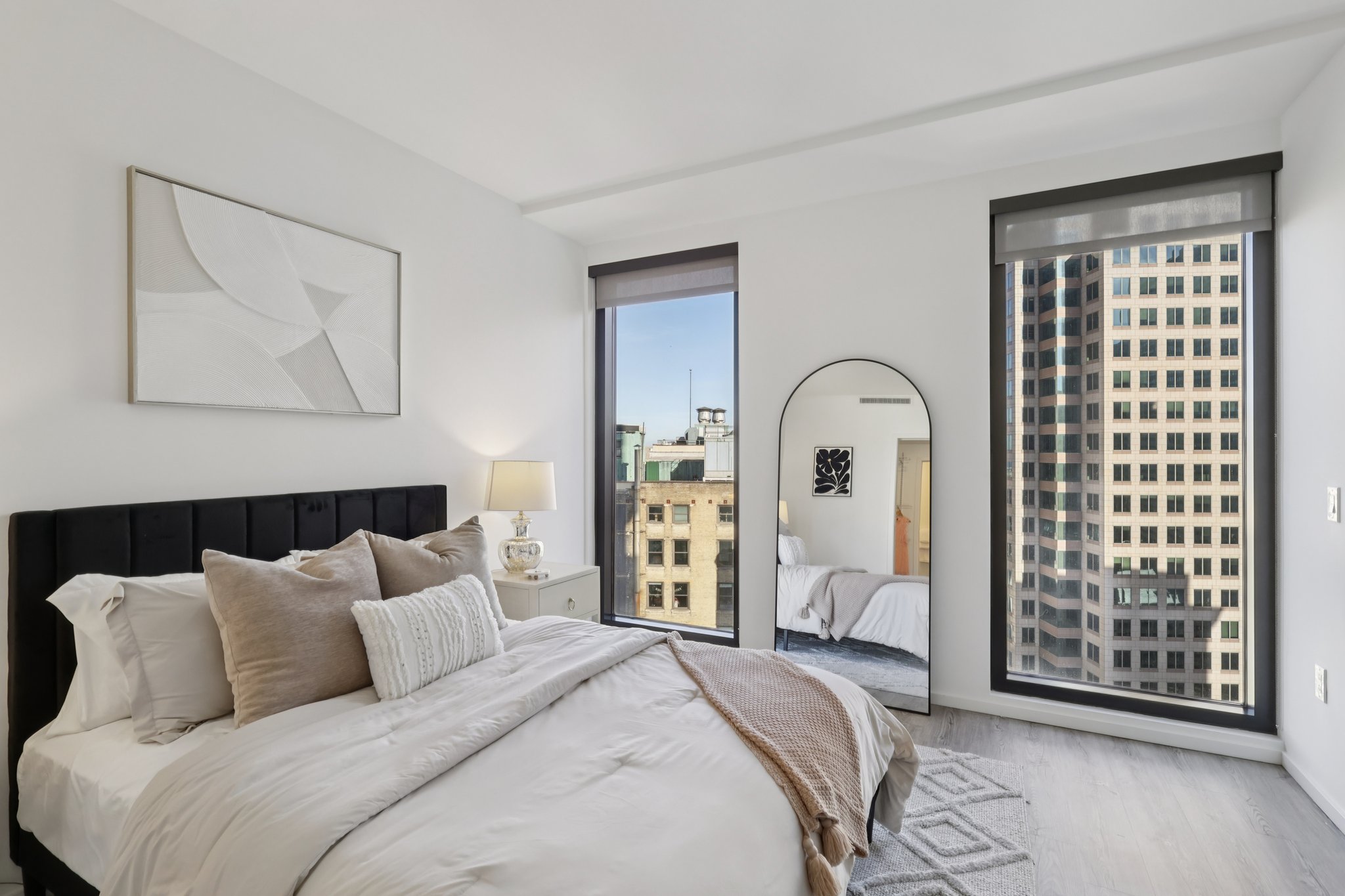 A modern bedroom featuring a black upholstered bed with white bedding and decorative pillows. There's a large mirror on one side and a window showcasing a city skyline. Soft lighting creates a cozy ambiance, accentuated by a minimalist wall art piece and a bedside lamp.