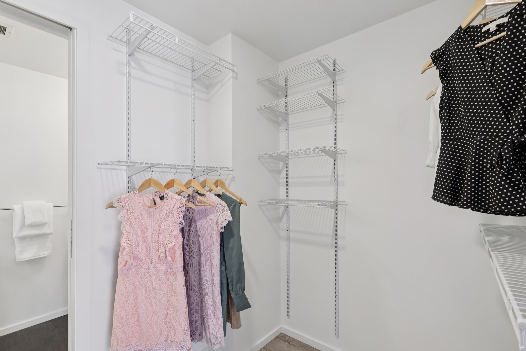 A well-organized closet featuring a variety of clothing items including pastel dresses and a black polka dot blouse. The closet has white wire shelves and hanging space on wooden hangers. The walls are painted white, and there is a bright, modern aesthetic.