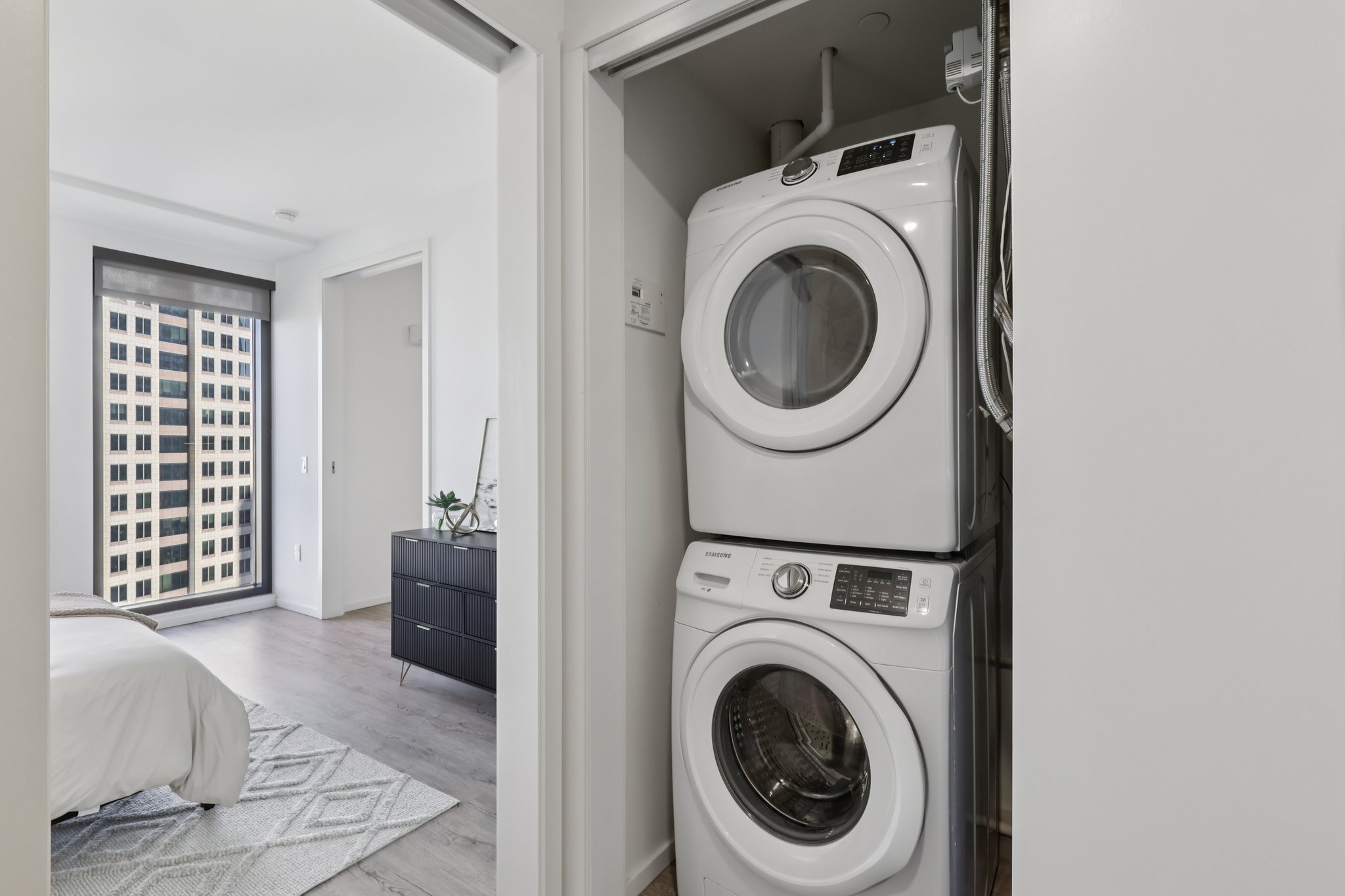 A compact laundry area features a stacked washer and dryer, located near a modern bedroom with large windows showing a city view. The room includes a stylish dresser against the wall and a light rug on the floor.