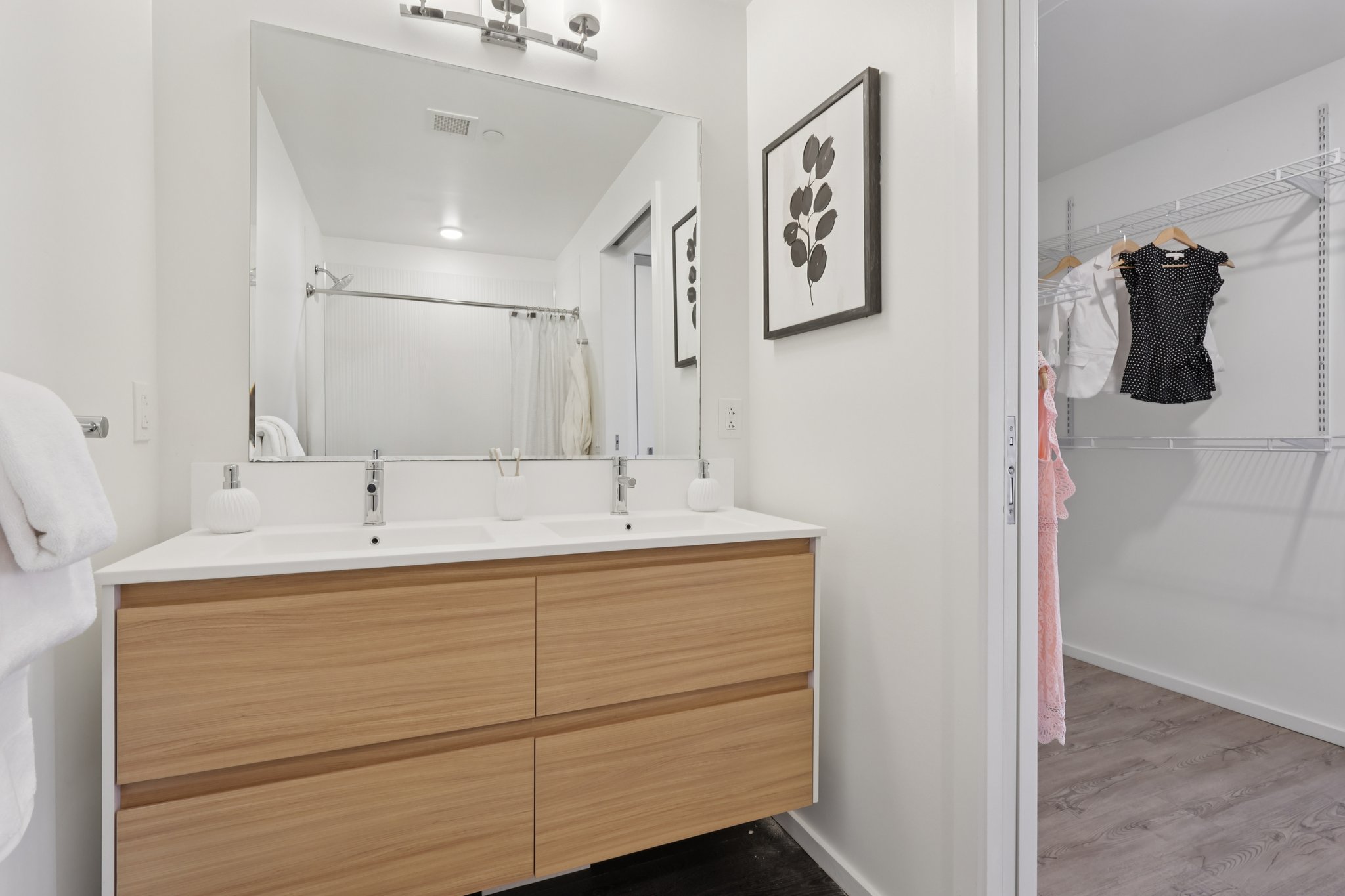 A modern bathroom featuring a double sink wooden vanity with two white soap dispensers, a large mirror above, and a bright, minimalist design. On the side, there is a small closet with hangers displaying clothes. The space is well-lit and has a clean aesthetic, with a light color palette.