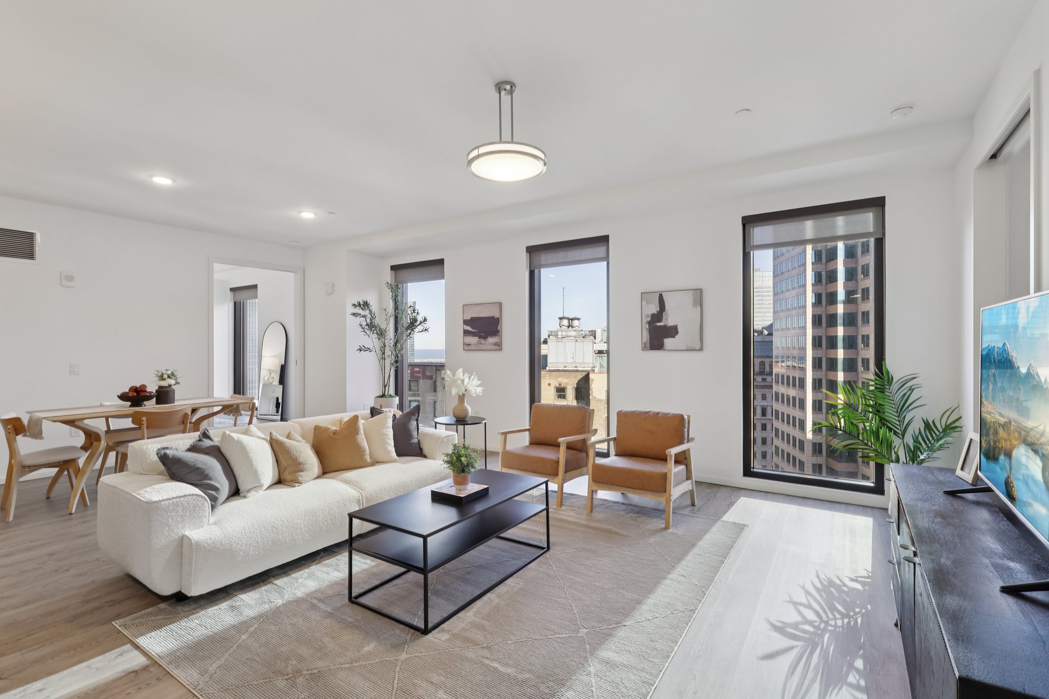 Spacious modern living room featuring a white sofa, two brown chairs, a black coffee table, and a TV stand. Large windows provide city views, while artwork adorns the walls. A dining table is visible in the background, creating an inviting atmosphere with natural light.