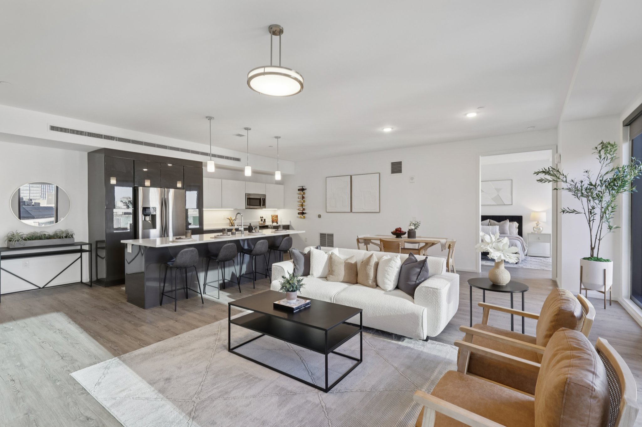 Modern open-concept living space featuring a stylish living room with a beige sofa, brown armchairs, and a black coffee table. The kitchen has dark cabinetry and a large island with seating. Large windows provide natural light, and minimalist decor adds a contemporary feel to the space.