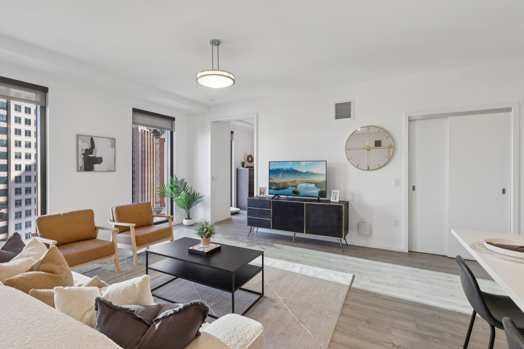 A modern, stylish living room featuring a light-colored sofa, an accent armchair, and a sleek black coffee table. A television mounted on the wall displays a scenic landscape. Large windows provide natural light, while a potted plant adds a touch of greenery to the space. The decor is minimalistic and contemporary.