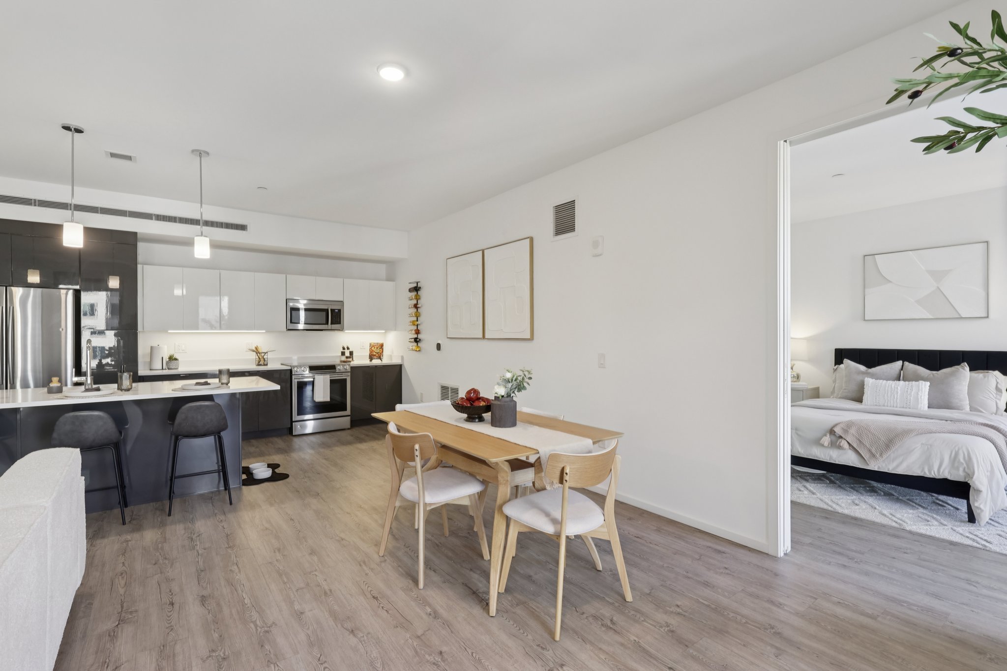 Modern open-concept living space featuring a kitchen with stainless steel appliances, a dining area with a wooden table and white chairs, and a cozy bedroom visible through an open door. The decor is minimalistic, with neutral colors and contemporary artwork. Natural light floods the room, creating a bright atmosphere.