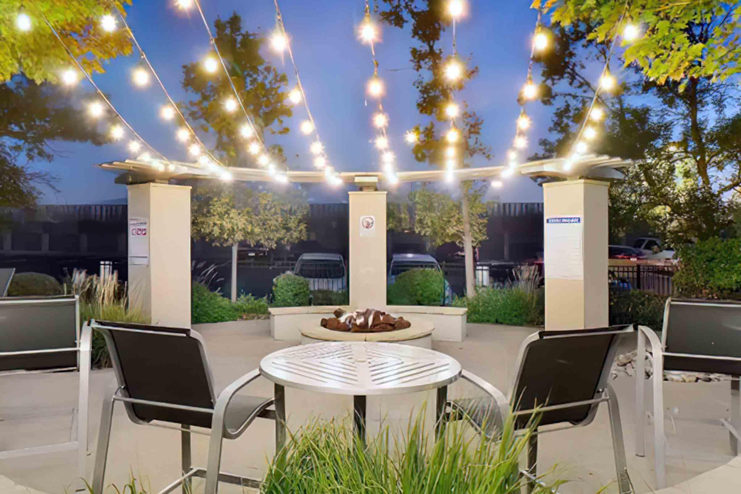A cozy outdoor seating area with two black chairs and a round table. Above, string lights illuminate the space, enhancing the ambiance. In the center, a fire pit is surrounded by potted plants and decorative stones. The setting is inviting, perfect for relaxing or gathering with friends in the evening.
