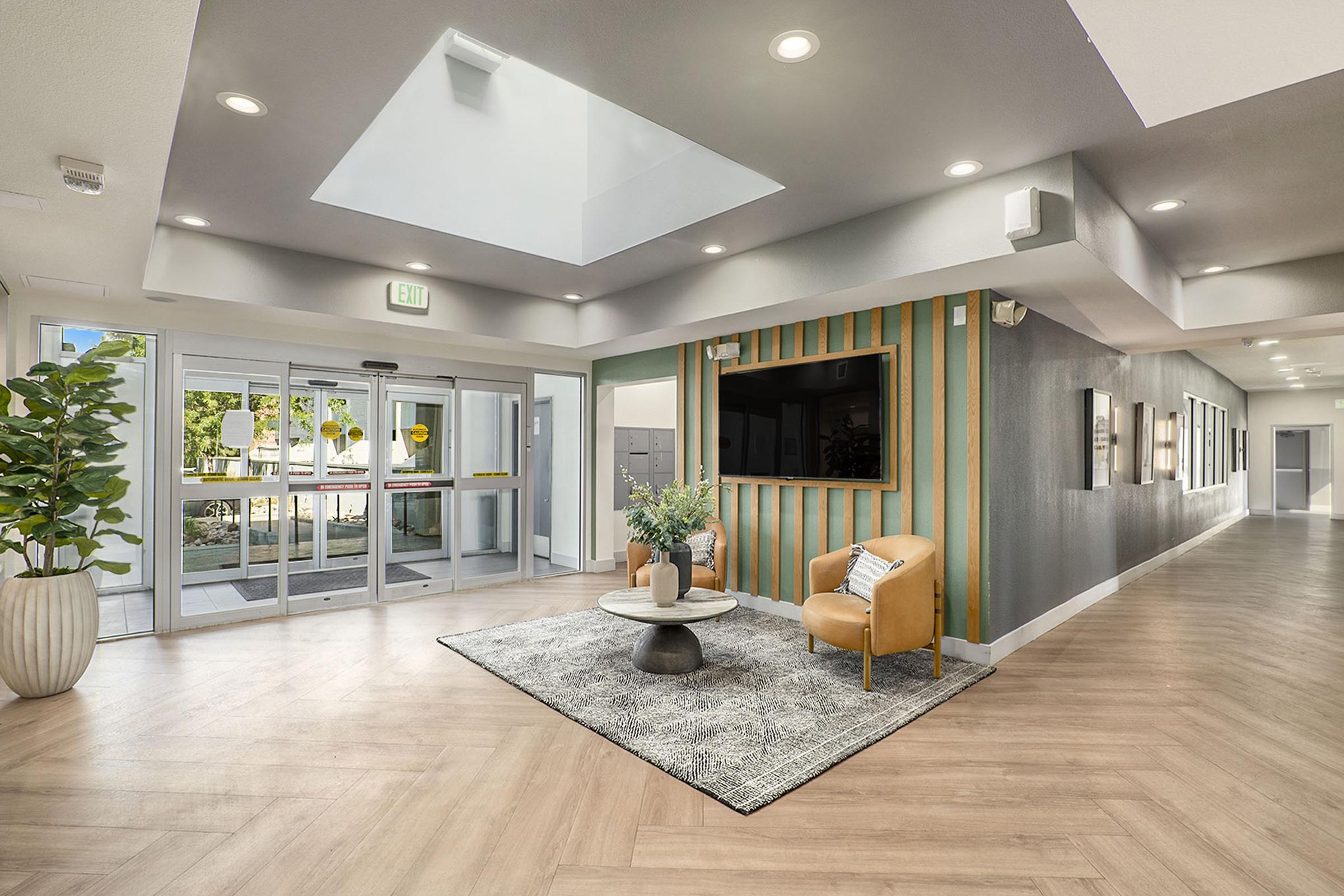 Bright and modern lobby featuring large glass doors, a decorative rug, a coffee table, and a stylish armchair. A potted plant adds a touch of greenery, while a wall-mounted TV is visible on the right side, surrounded by green and wooden paneling. Natural light enters through a skylight above.