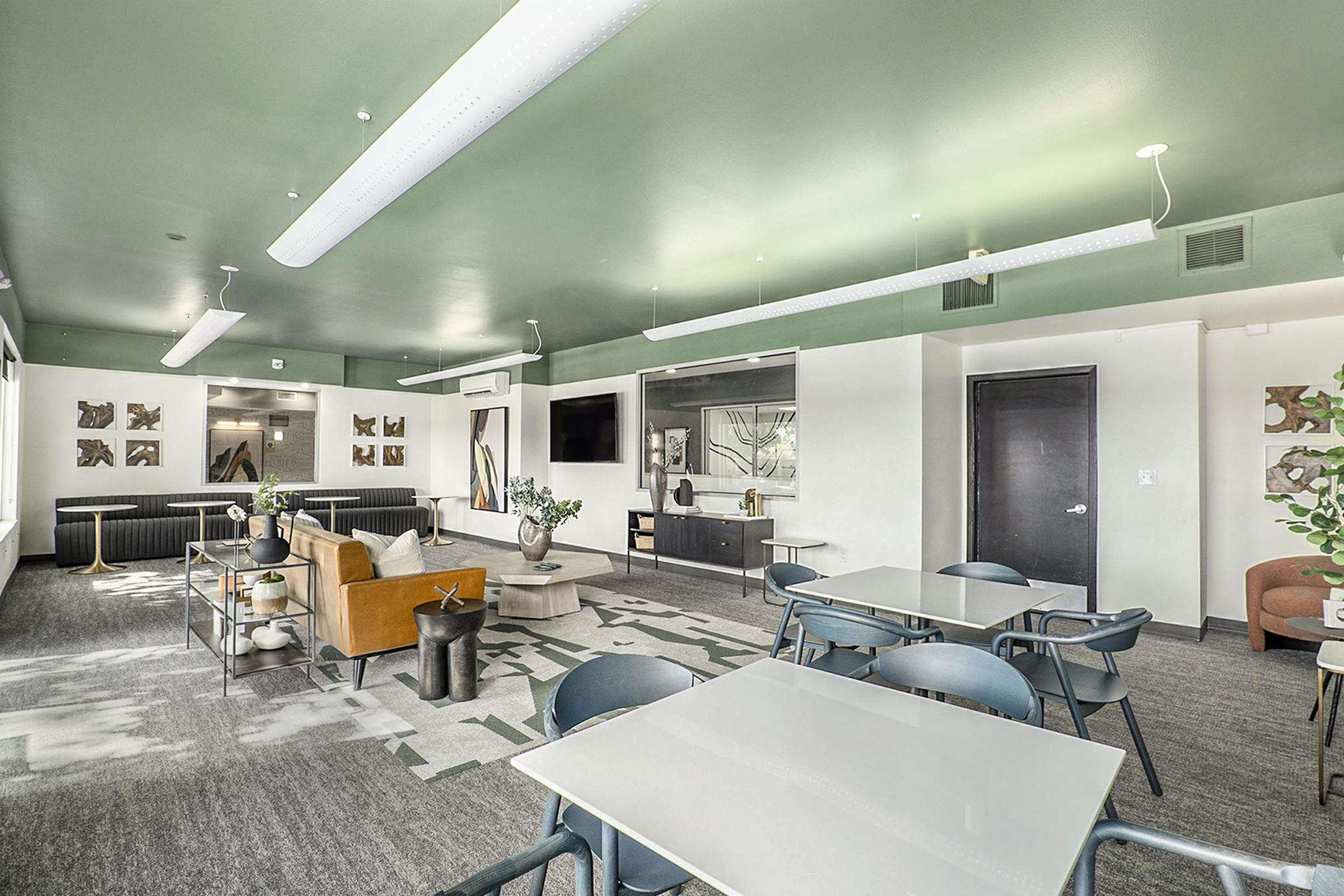 Bright and modern common area with green ceiling, featuring several tables and chairs, a comfortable seating arrangement with an orange chair, and a large wall-mounted TV. Decorative plants and artwork enhance the space, promoting a welcoming atmosphere for gatherings or relaxation.