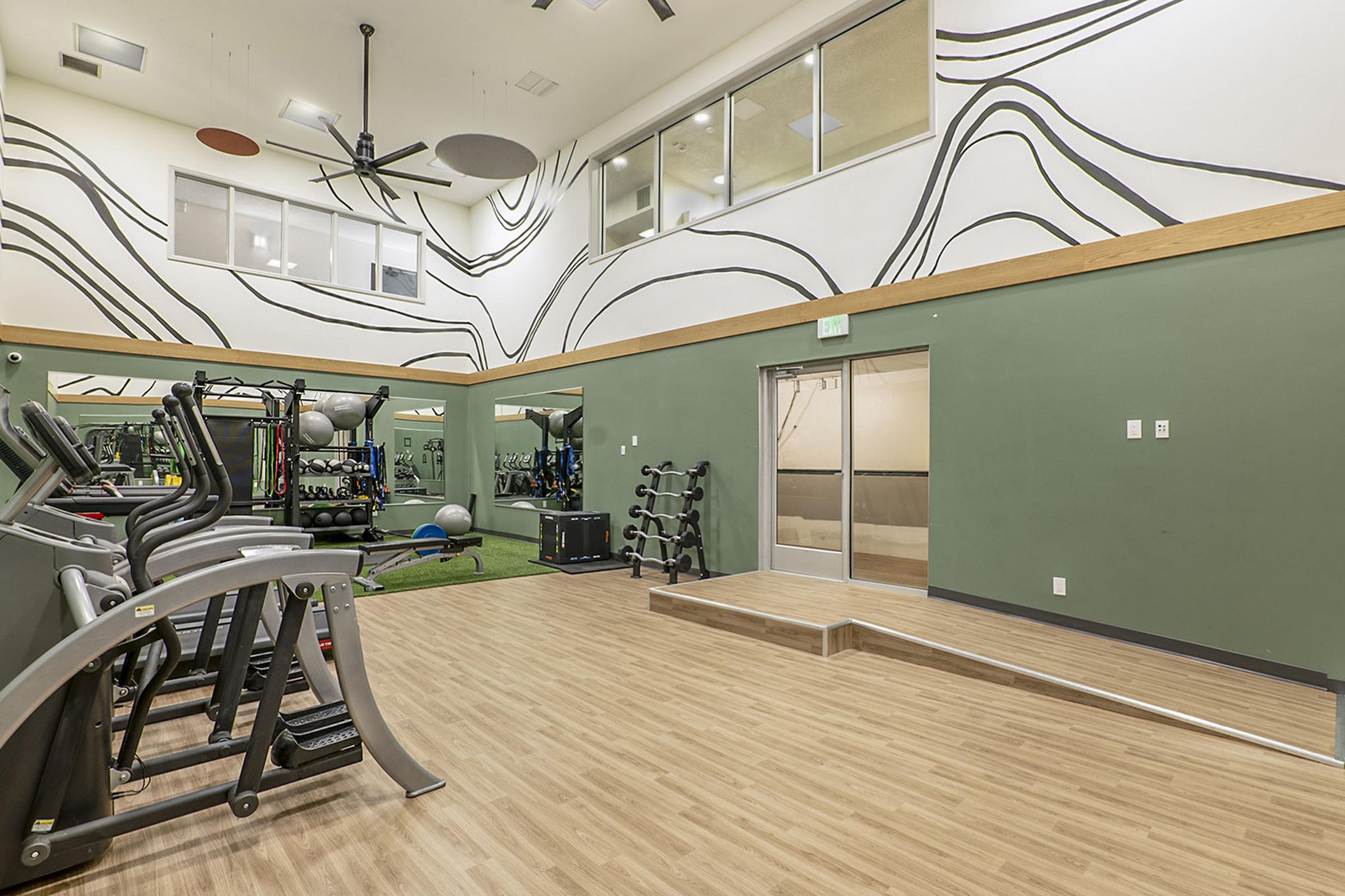 Interior of a modern gym featuring exercise equipment, including treadmills and weights. The walls are decorated with abstract design elements. Large windows provide natural light, and there is a small raised platform with an entrance. The flooring is wood laminate, creating a warm atmosphere.