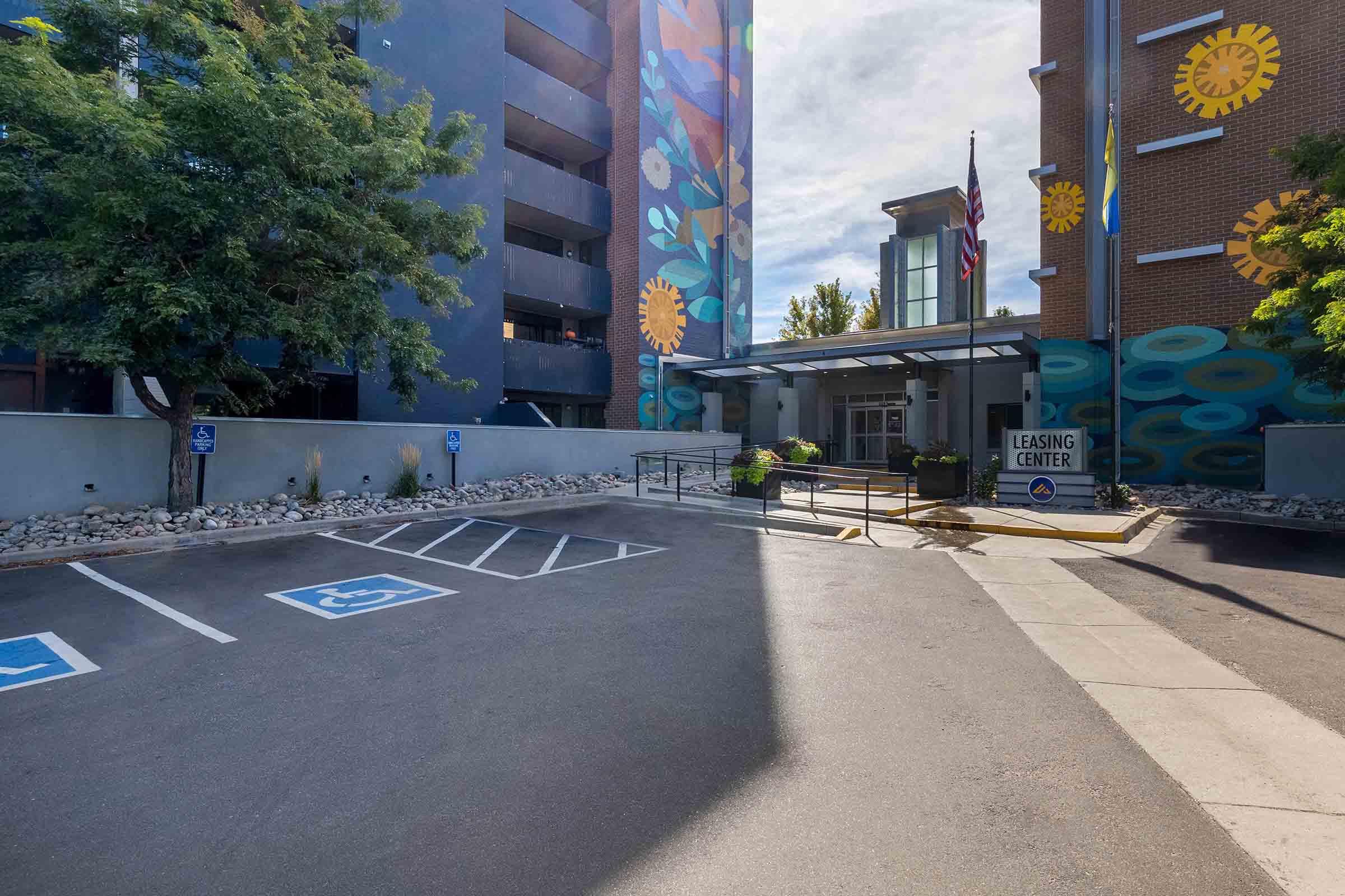 A leasing center building with colorful murals on the side and flags out front. There is a parking area with marked handicapped spaces and a pathway leading to the entrance. The scene is well-lit with trees and landscaping visible.