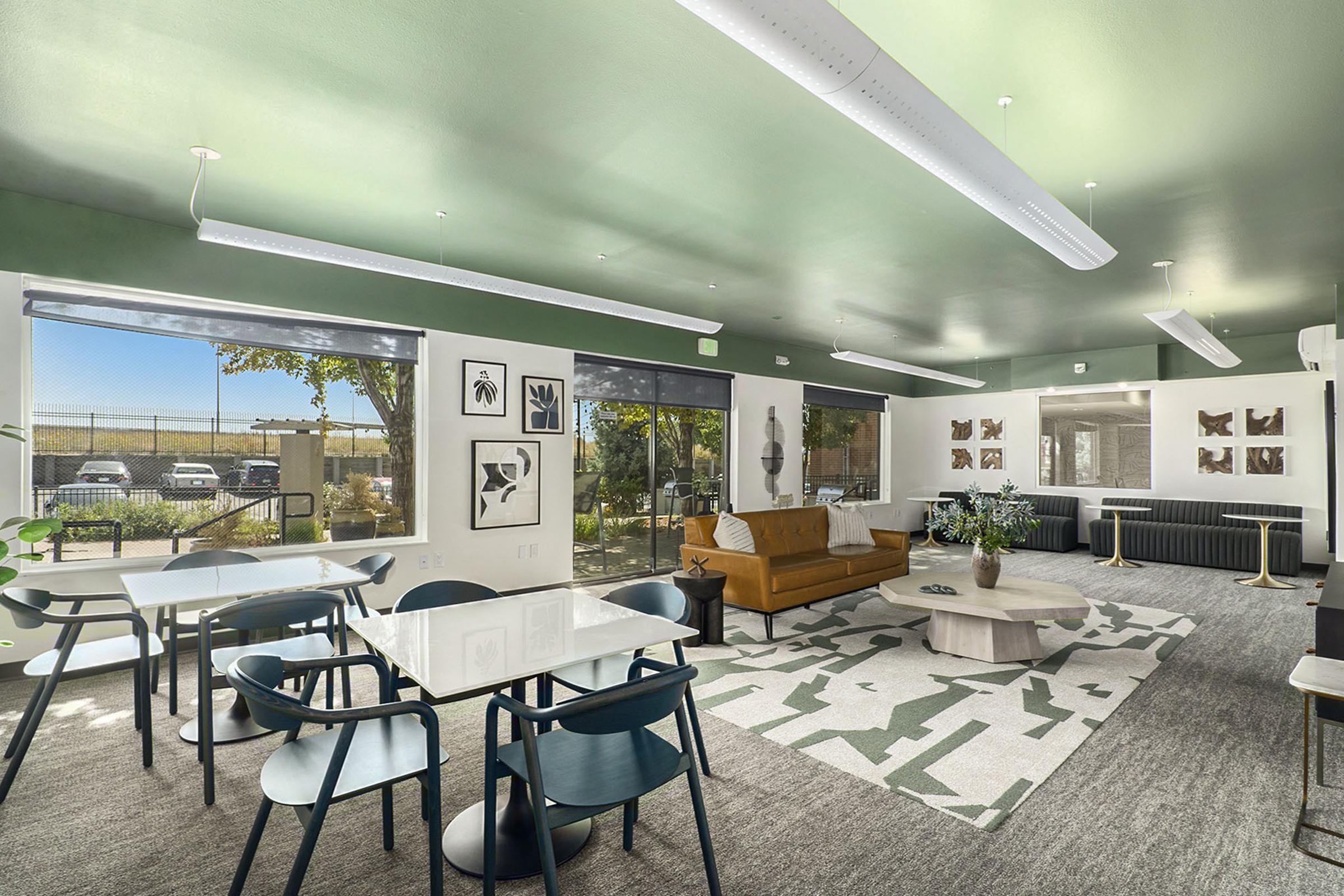 Modern, well-lit communal room featuring a cozy seating area with a brown sofa, a coffee table, and artwork on the walls. There are several dining tables and chairs, large windows allowing natural light, and a view of greenery outside. The ceiling is painted green, enhancing the contemporary design.