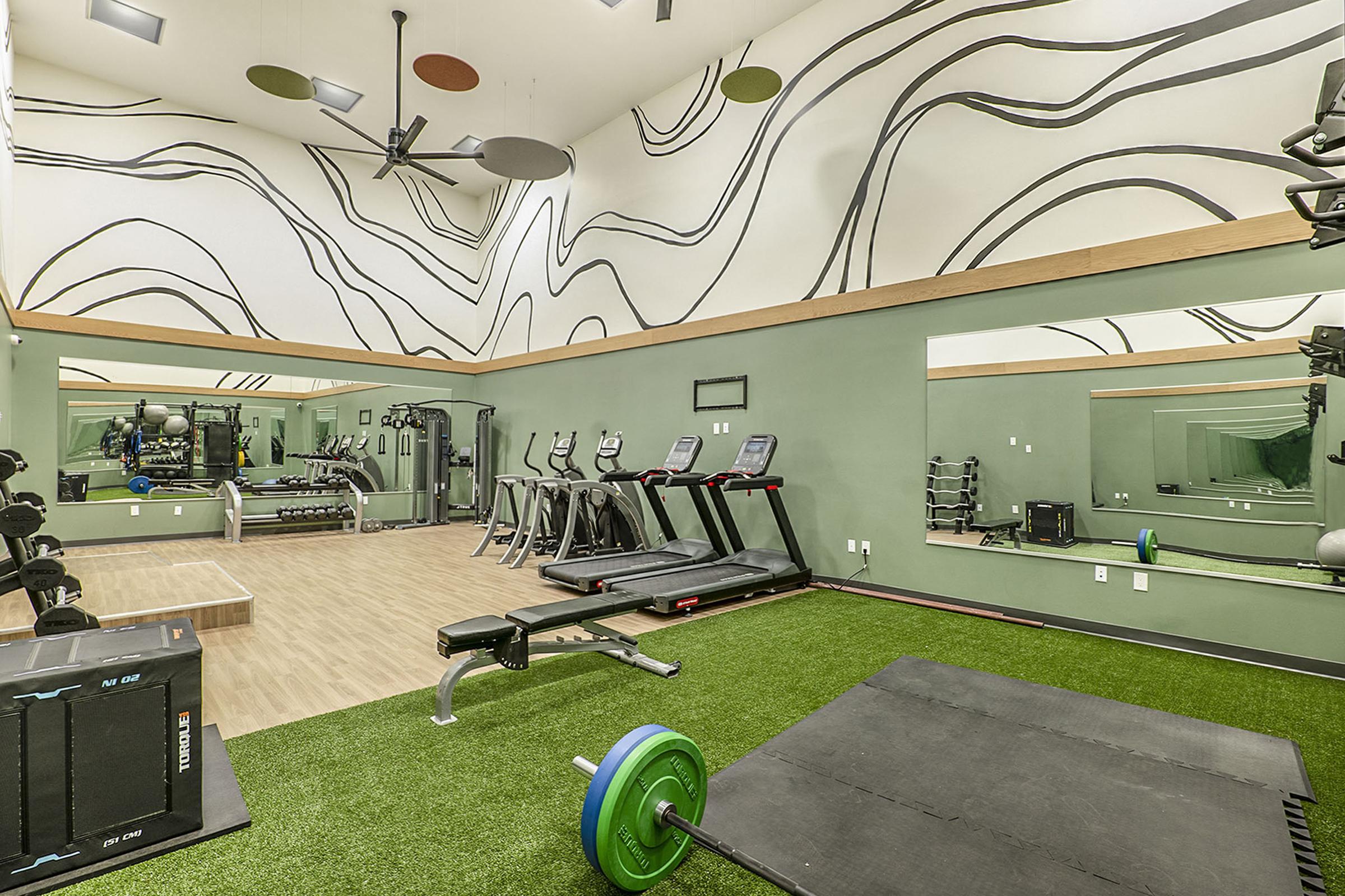 A modern gym interior featuring workout equipment including treadmills, free weights, and a weight bench. The space has a bright, open design with a high ceiling, green walls, and abstract patterned wall art. There is a mirrored wall reflecting the equipment and a turf area for exercises.