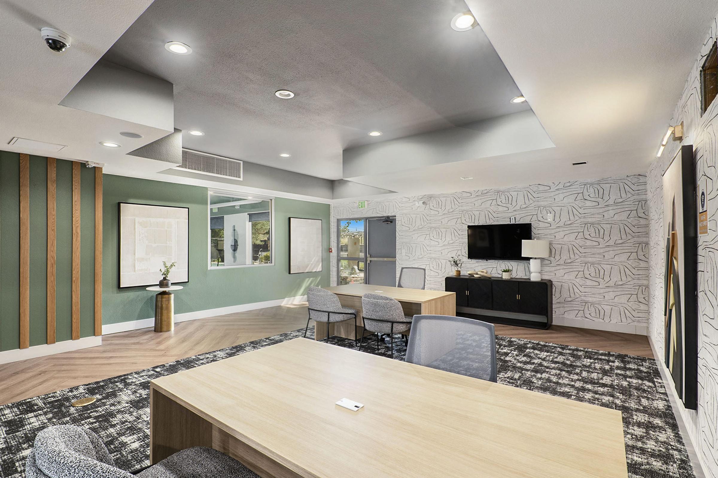 A modern office interior featuring a green accent wall, stylish artwork, a large wooden table with chairs, and a wall-mounted TV. The room has a cozy ambiance with layered lighting, patterned flooring, and decorative elements, making it suitable for meetings or collaborative work.