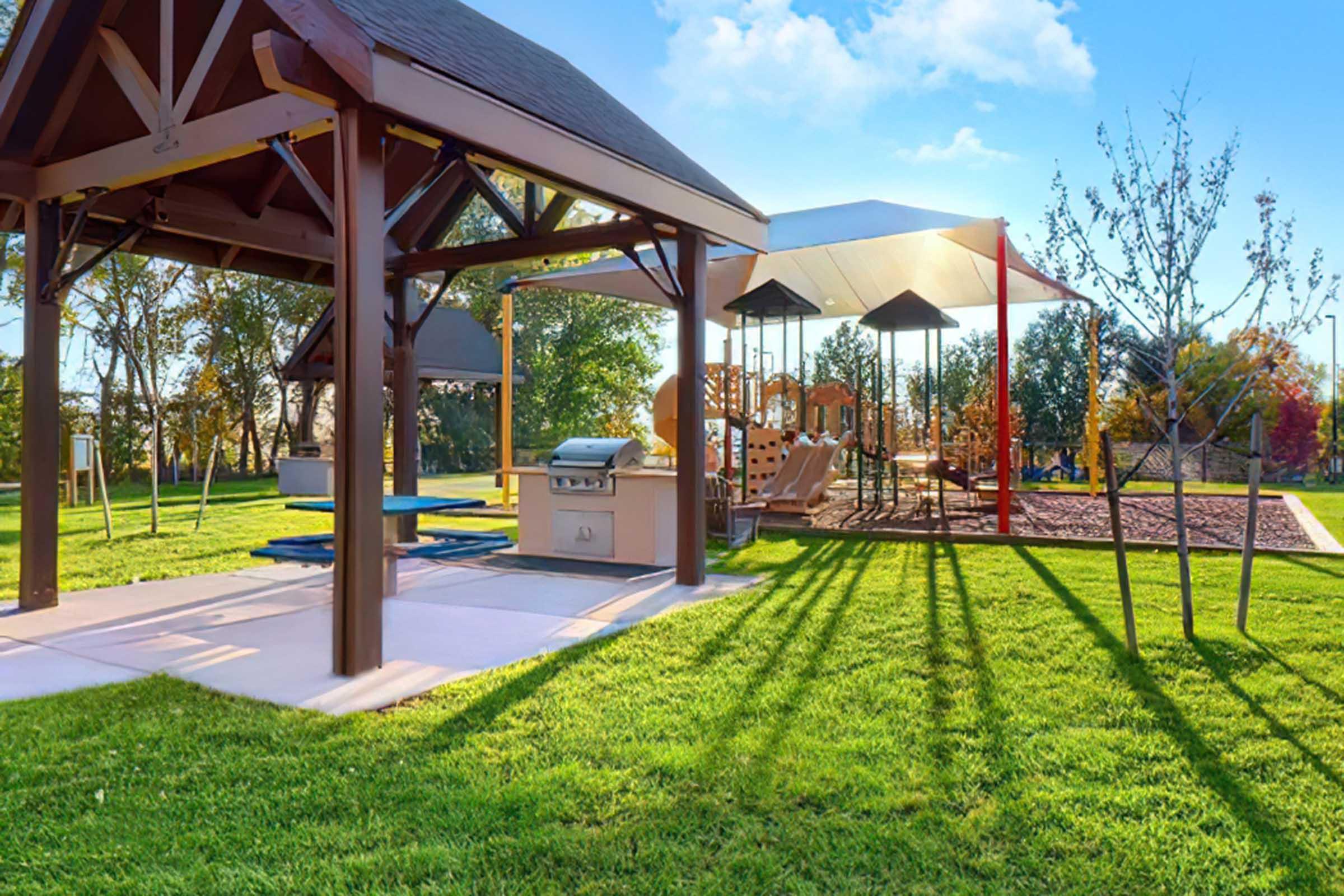 A sunny park scene featuring a picnic area with a grill and blue tables under a wooden pavilion, alongside a playground with swings and slides. Lush green grass surrounds the area, and several young trees are visible, creating a welcoming outdoor space for families and children.