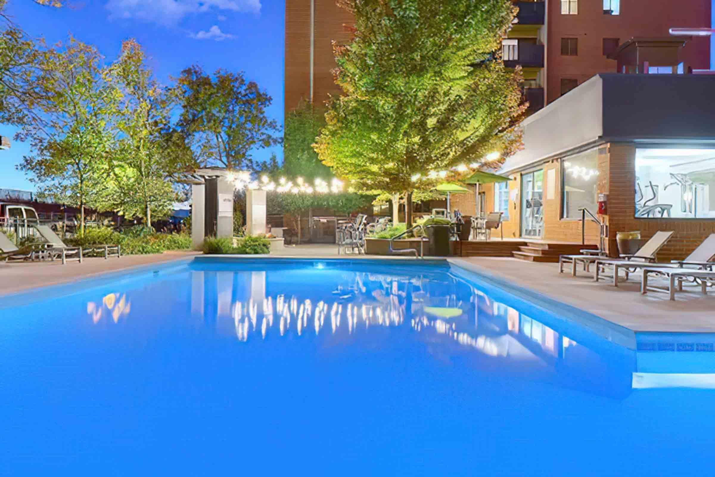 A tranquil swimming pool illuminated by string lights at dusk, surrounded by lounge chairs and green trees. The water reflects the colorful sky, creating a serene and inviting atmosphere for relaxation and evening enjoyment.