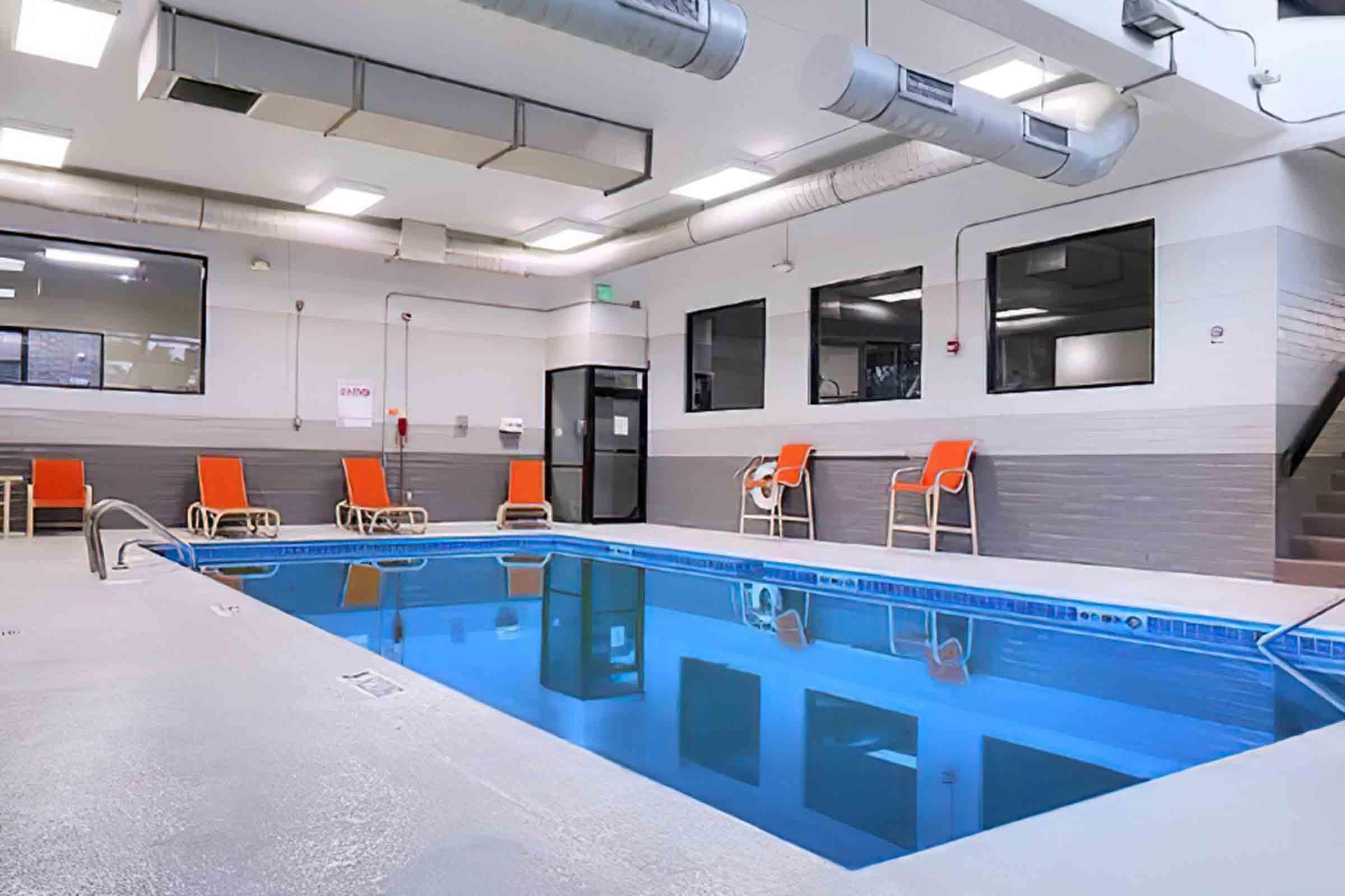 Indoor swimming pool area featuring clear blue water, surrounded by several orange lounge chairs. The space has large windows allowing natural light in, and a staircase leads to another level. The design is modern and well-lit, creating a welcoming atmosphere for relaxation and leisure.