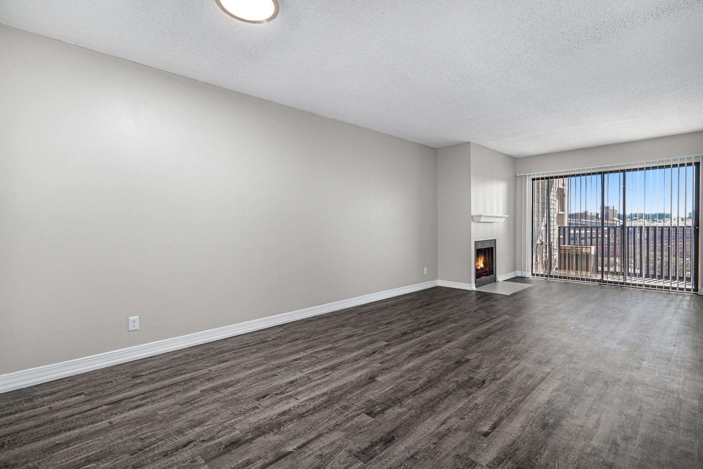 A spacious, modern living room with light-colored walls and dark wood-style flooring. There is a fireplace to the right and large sliding glass doors leading to a balcony. The room is bright with natural light, showcasing an open layout, perfect for relaxation or gatherings.
