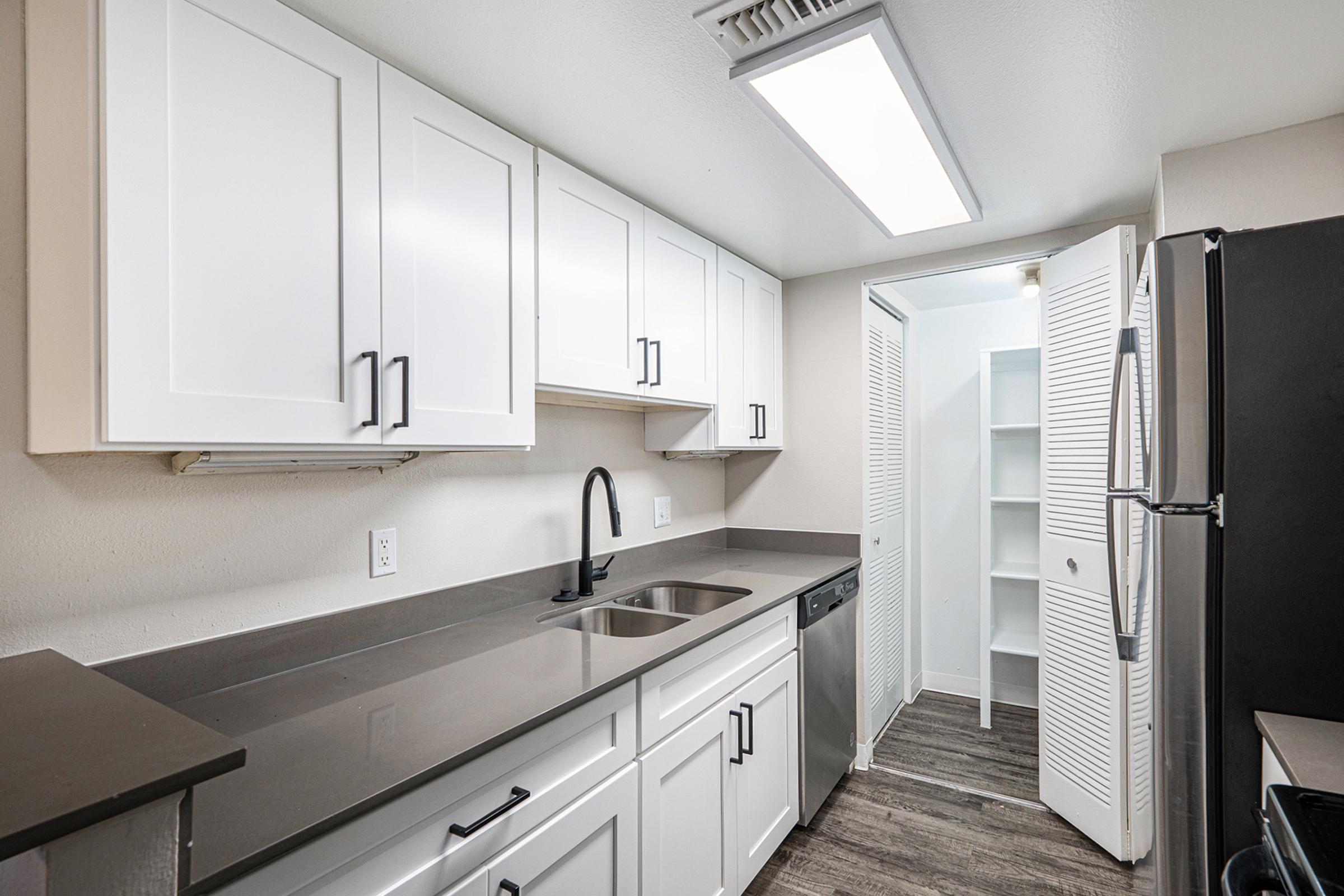 Modern kitchen featuring white cabinets with black handles, a stainless steel sink, dishwasher, and dark countertops. A refrigerator is on the right, and there is a pantry or storage closet visible in the background with bi-fold doors. The space is well-lit with overhead lighting.