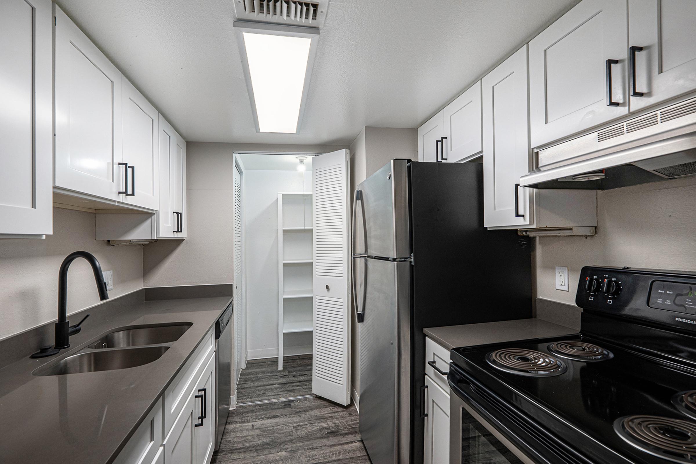 A modern kitchen featuring white cabinets, a dark countertop, and stainless steel appliances. The space includes a black refrigerator, an electric stove, and a double sink. A pantry with shelves is visible through an open door. The flooring is a wood-like laminate, enhancing the contemporary feel of the kitchen.