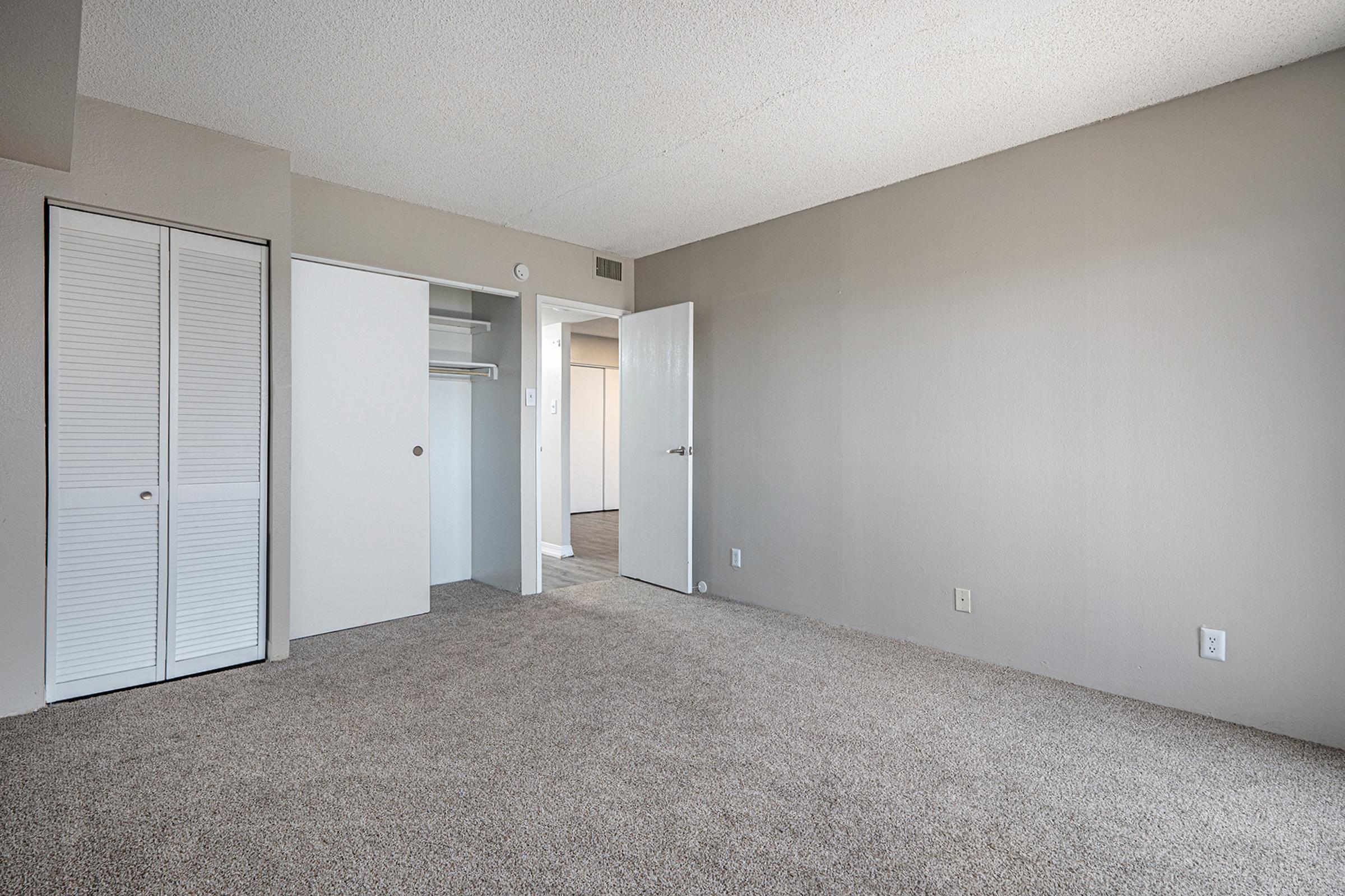 A spacious, empty bedroom featuring light grey walls and carpet. The room has a white closet with sliding doors on the left and an open door leading to another space on the right. Natural light brightens the area, highlighting its simple and minimalist design.
