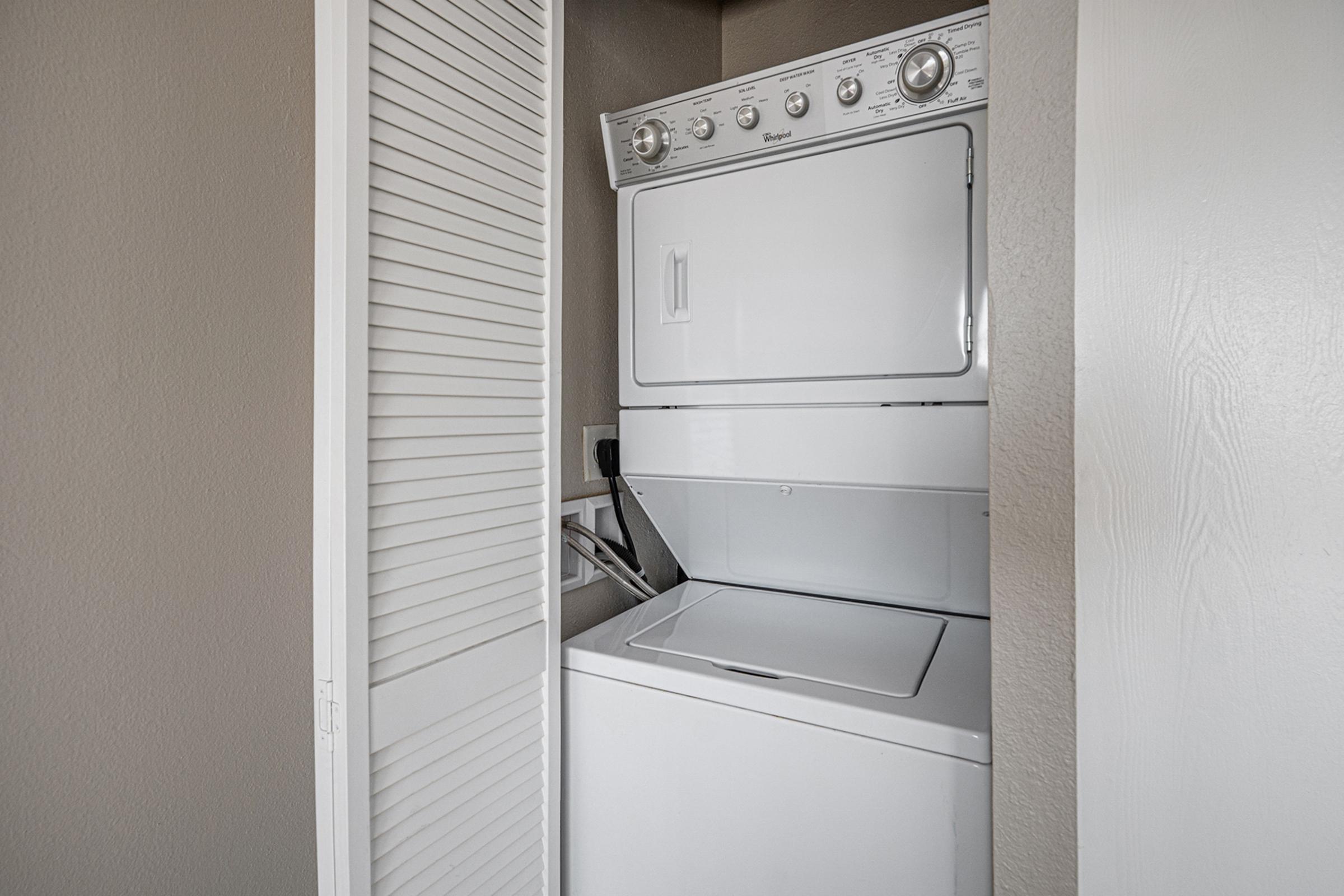 A stacked washer and dryer unit located in a small laundry space with beige walls and a partially open closet door, providing a functional and compact design for laundry chores.