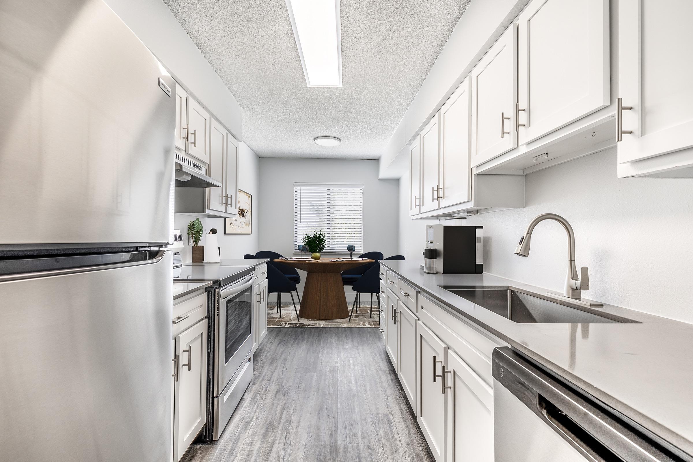 Modern kitchen featuring stainless steel appliances, including a refrigerator and oven, white cabinetry, and a sleek countertop. A dining area with a wooden table and black chairs is visible through the window, illuminated by natural light. The space is bright and well-organized.