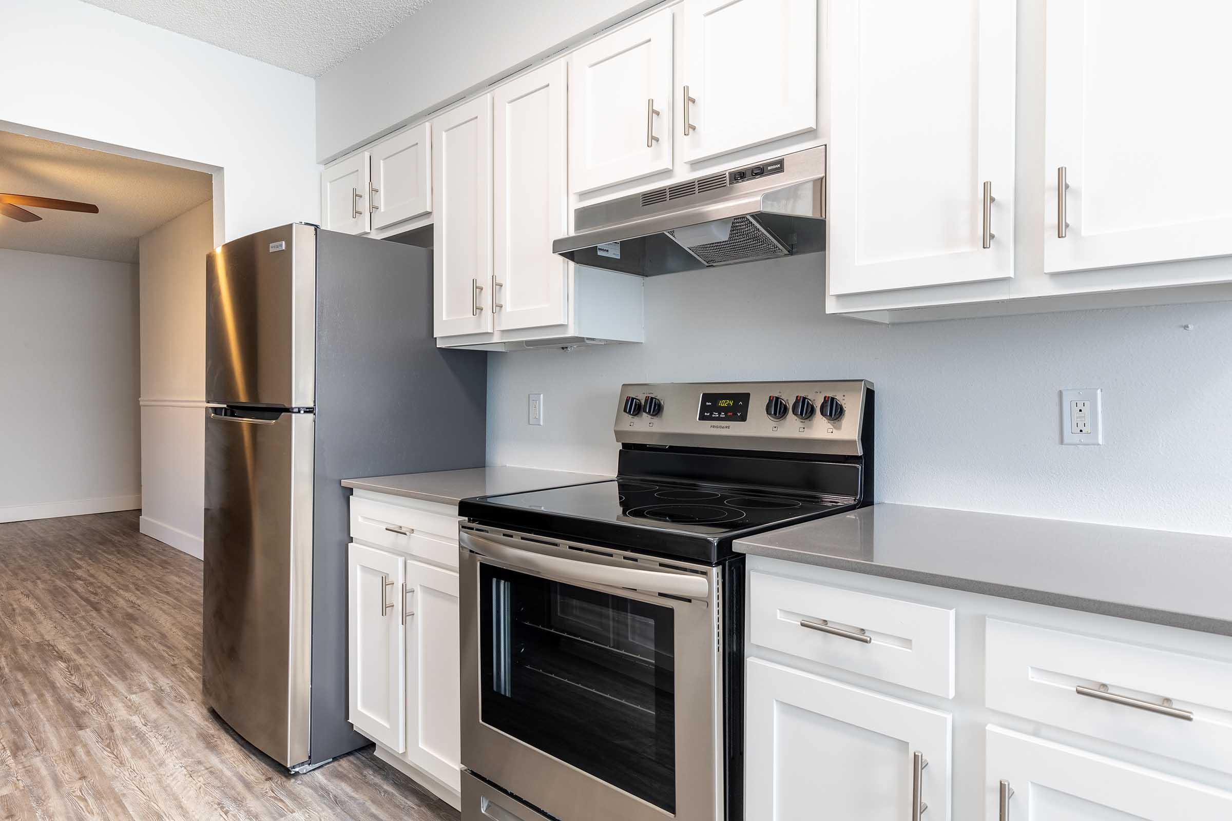 Modern kitchen featuring stainless steel appliances including a refrigerator and stove, white cabinetry, and a sleek countertop. The space has light-colored walls and a wood-like floor, creating a clean and contemporary look. A ceiling fan is visible in the adjacent room.