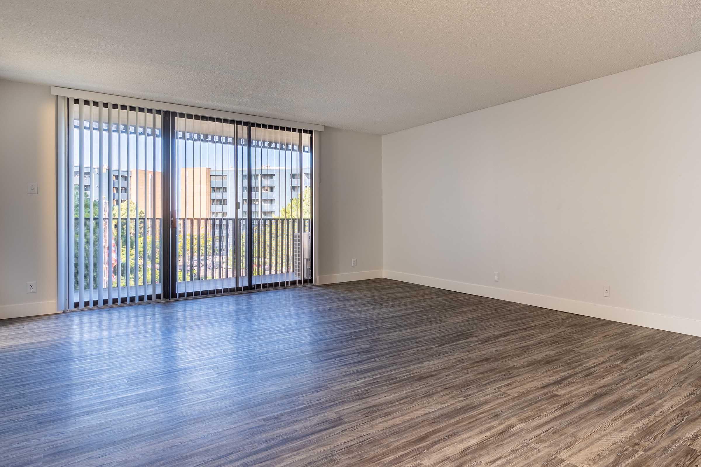 A spacious, empty room with large sliding glass doors featuring vertical blinds that open to a balcony. The floor is covered with light brown laminate flooring, and the walls are painted in neutral tones. Sunlight streams in, highlighting the openness of the space.