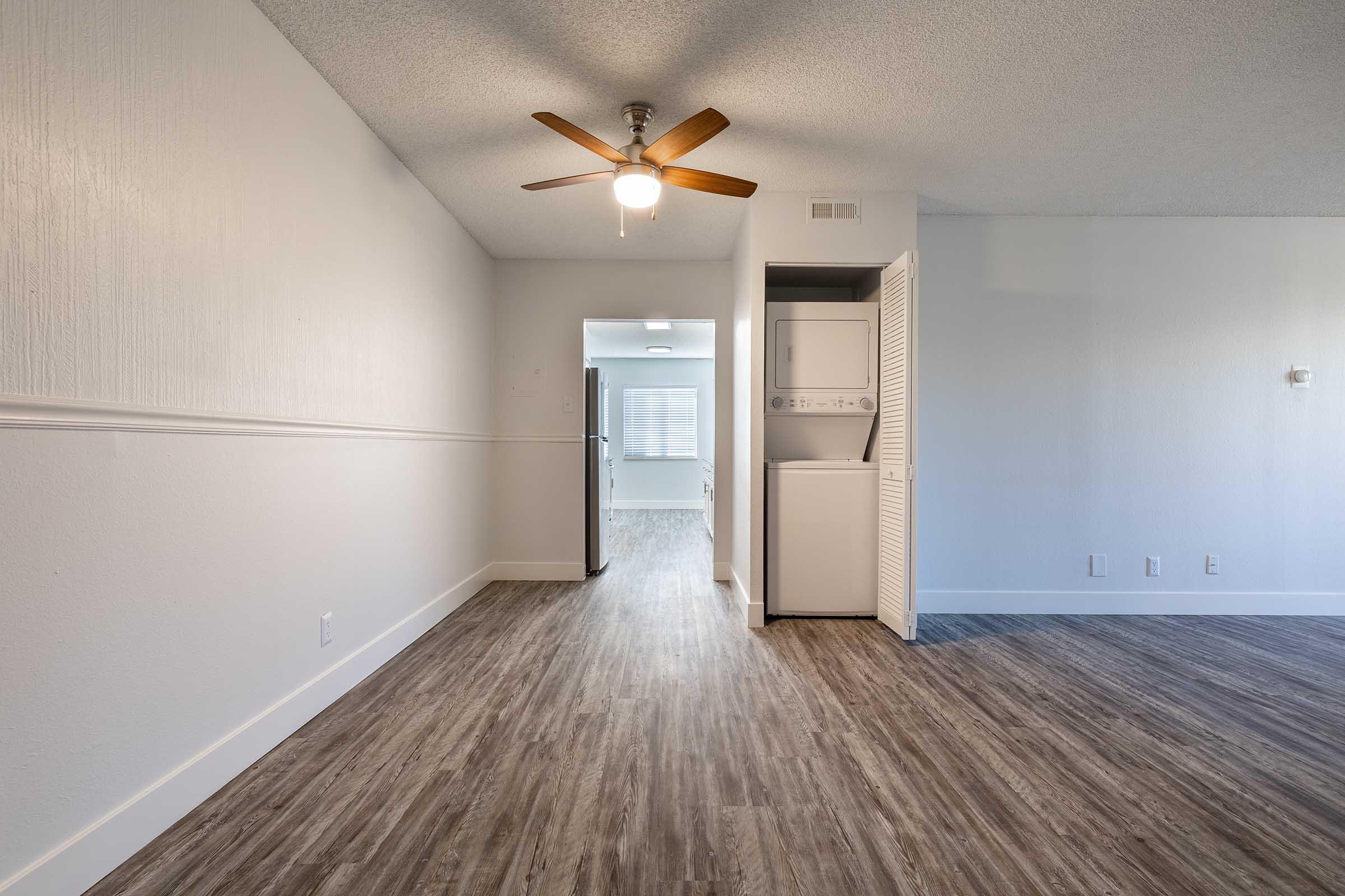 A spacious, well-lit interior of an apartment featuring a ceiling fan, laminate flooring, and a laundry unit in a closet. The room has neutral-colored walls and leads to a modern kitchen area visible in the background.