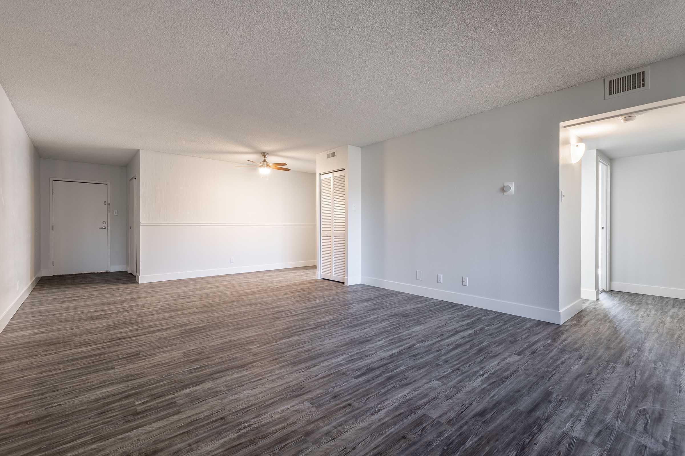 Spacious interior of an empty room featuring light-colored walls, a ceiling fan, and modern laminate flooring. A doorway leads to another space, while a door on the left suggests access to another area. Natural light enhances the clean, open atmosphere, ideal for customization.