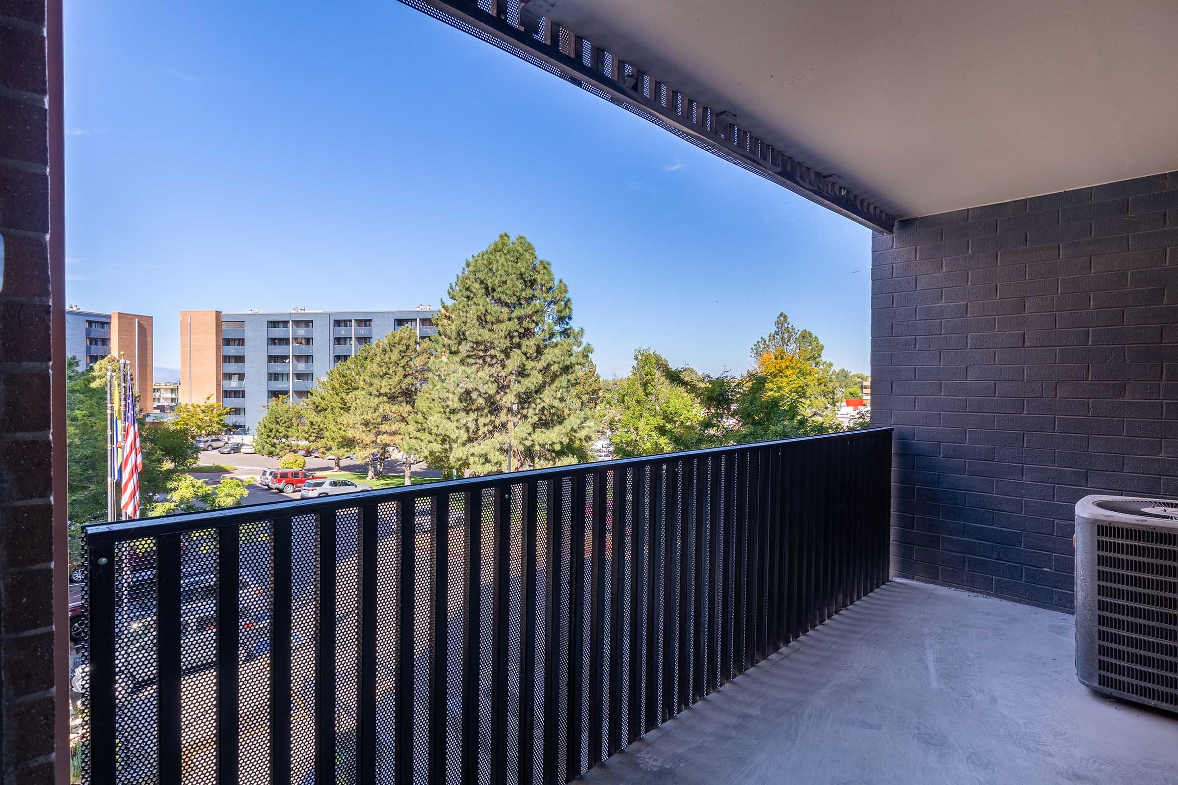 View from a balcony featuring a black railing, overlooking a tree-lined area with apartment buildings in the distance. The sky is clear and blue, and there's an outdoor air conditioning unit positioned on the right side of the balcony. The scene conveys a suburban atmosphere with greenery.