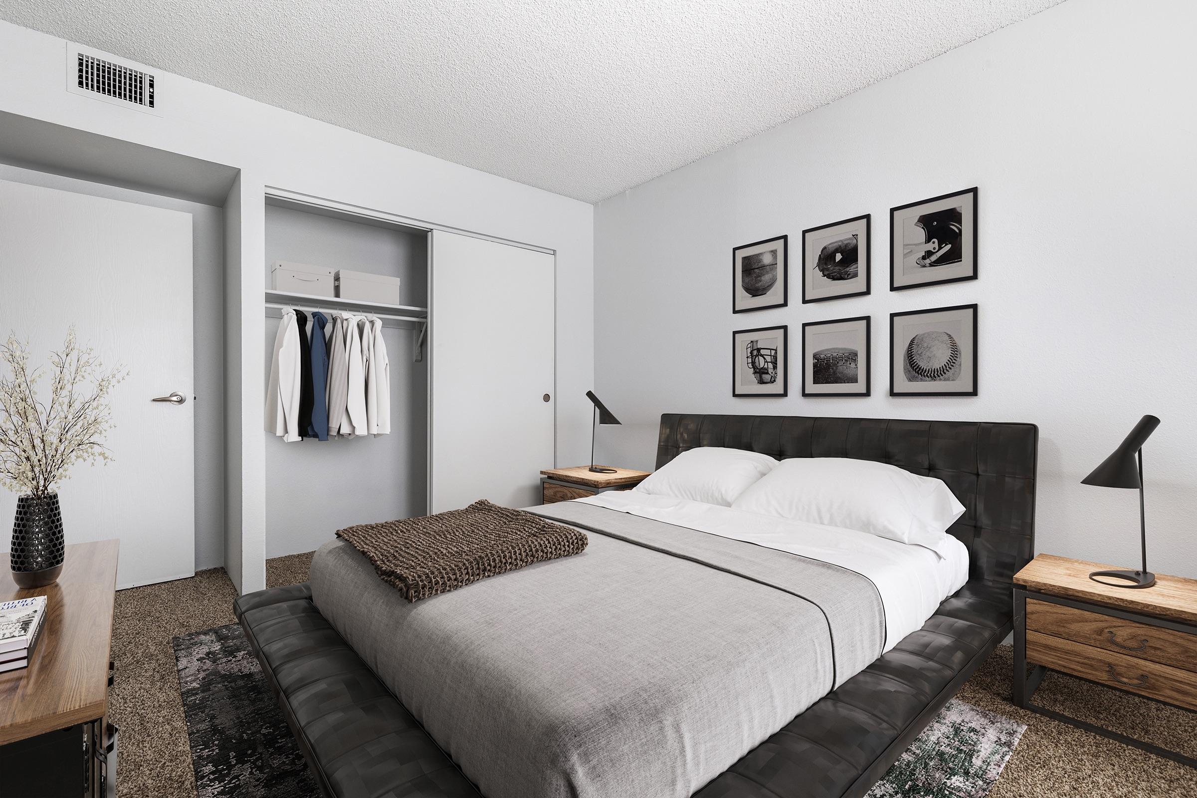 A modern and minimalistic bedroom featuring a neatly made bed with gray bedding and a brown throw blanket. On either side of the bed, there are wooden nightstands with lamps. A closet is visible with hanging clothes, and four framed black-and-white photographs adorn the wall above the bed. The room has a neutral color scheme and contemporary decor.
