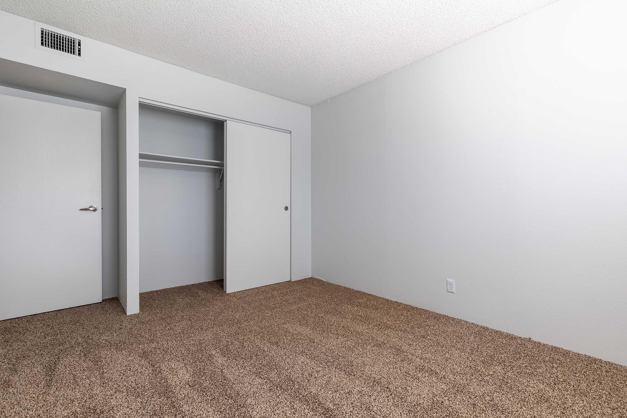 A vacant, softly lit room featuring beige carpet flooring. The walls are painted in a light gray color. There is a closed white closet door on the left, and a plain white door leading to another area is visible. The room has a minimalist and clean appearance, hinting at potential for personalization.