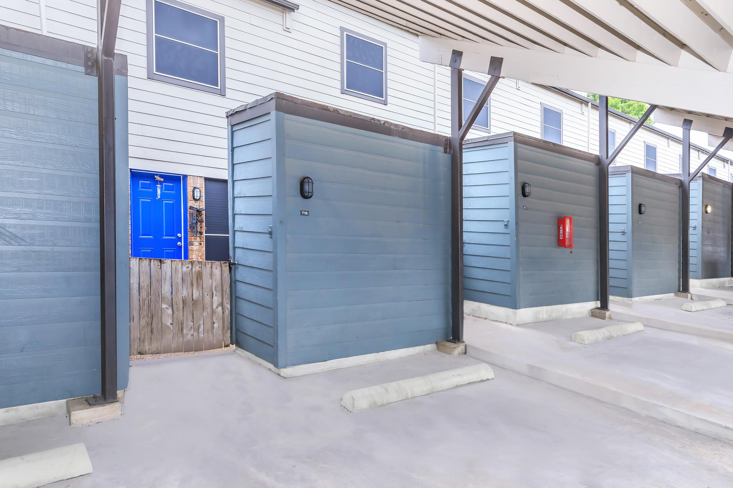A row of storage units in a covered area, with one unit featuring a bright blue door. The background includes a building with white siding and multiple windows. There is a wooden fence partially visible beside the blue door, and a red fire extinguisher is mounted on one of the storage units.