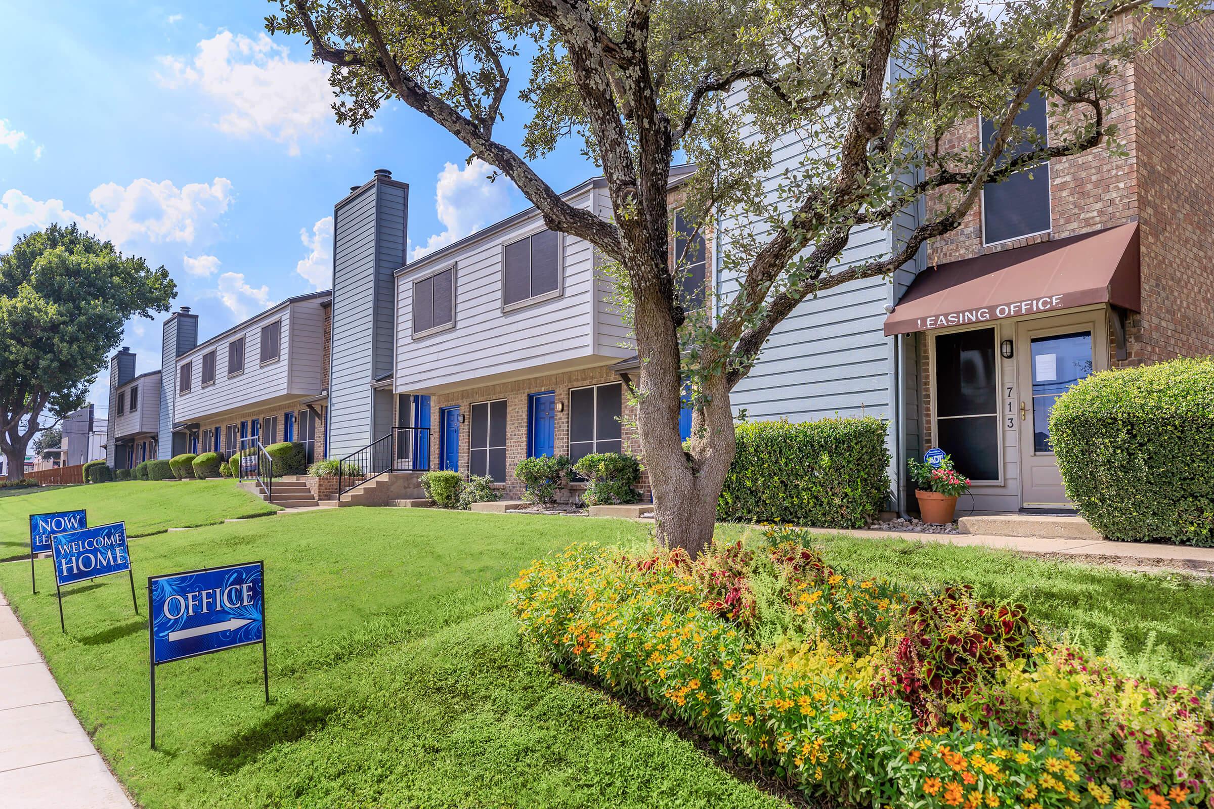 A well-maintained apartment complex featuring multiple buildings with a leasing office area. Lush green lawns and colorful flower beds are visible in the foreground, along with signage indicating "Now Leasing" and "Office." The sky is clear with a few clouds.