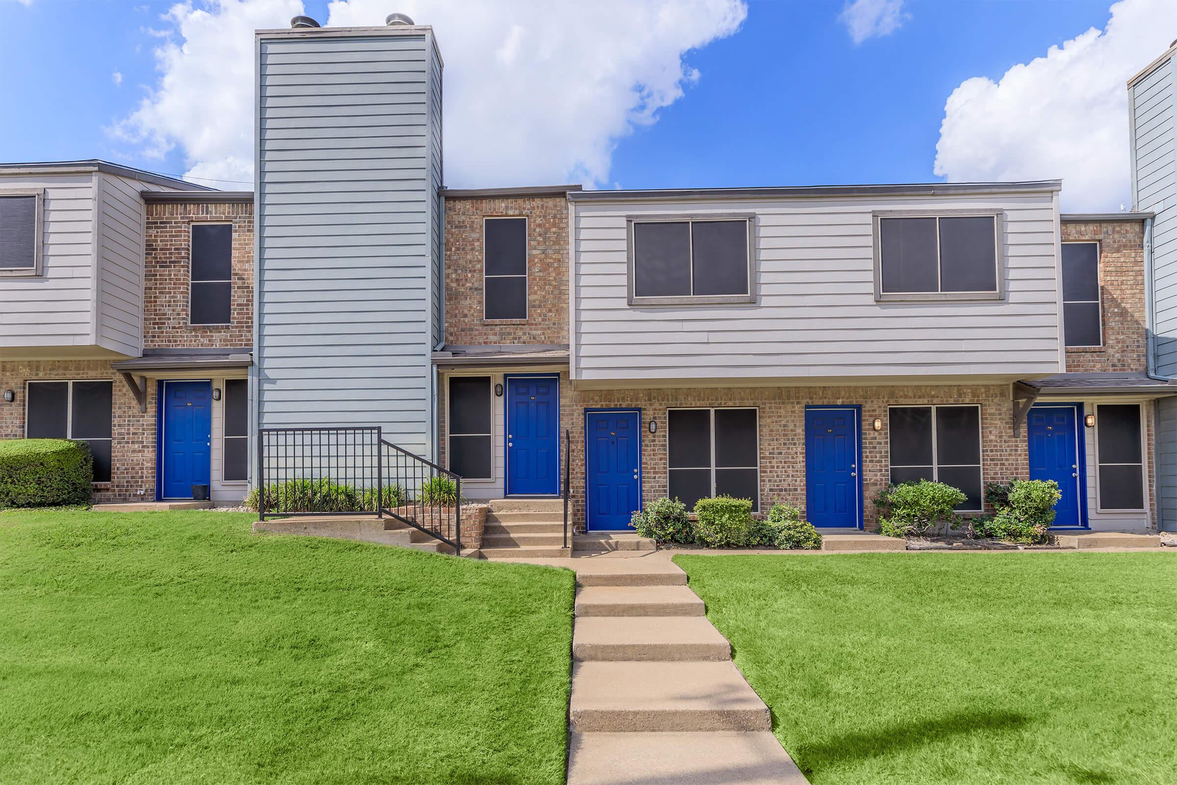 A row of townhouses featuring gray siding and blue doors. The buildings are set on a well-manicured lawn with neat landscaping. There are concrete pathways leading to the entrances, and a blue sky with some clouds is visible in the background.